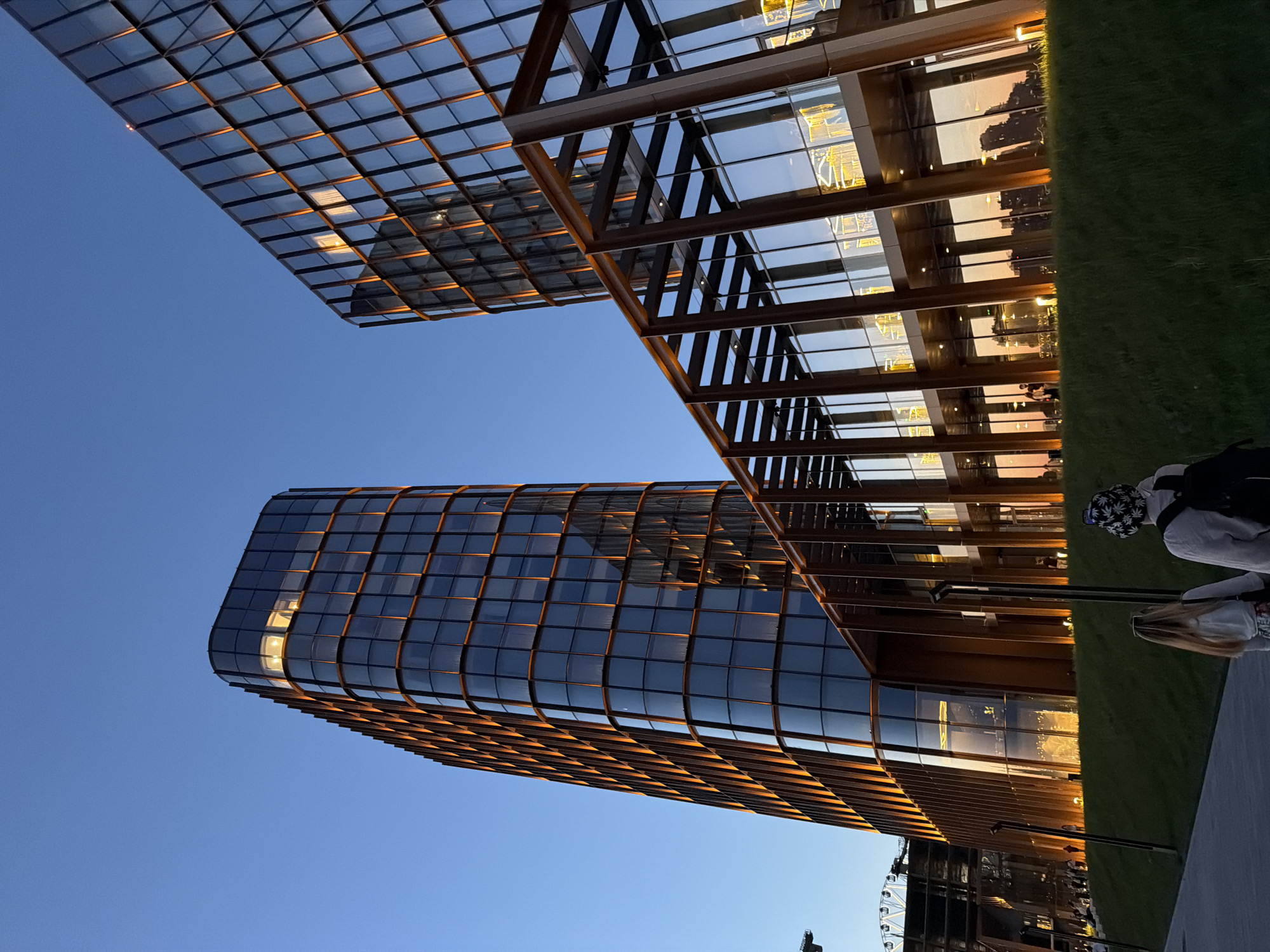 The image depicts a modern architectural scene in an unidentified urban location. The focus is on a tall, curved building with a grid-like glass facade, which reflects the sky and light. Adjacent to this building is an open structure with exposed beams and glass panels, possibly a covered walkway or architectural element.
In the background, hints of greenery and possibly a Ferris wheel are visible. Two people are partially visible on the right side of the image, one wearing a hat and a backpack. The lighting suggests it is either sunrise or sunset, casting warm tones on the buildings and sky. The environment appears to be a city or urban area.