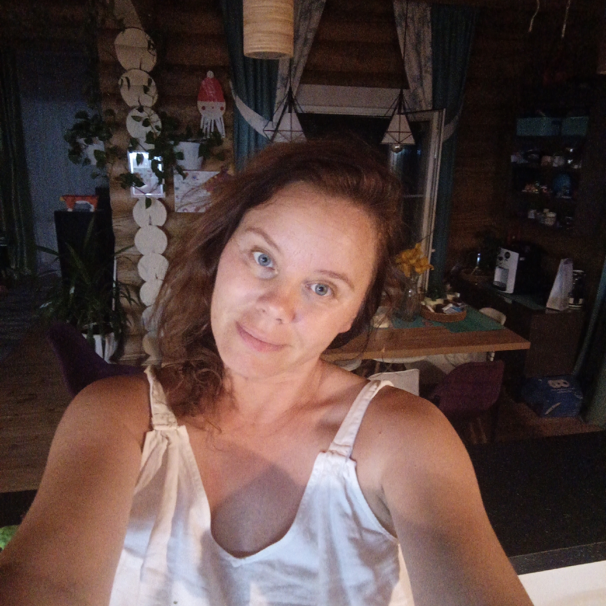 A medium shot captures a woman with auburn hair and blue eyes in what appears to be a rustic interior, possibly a log cabin. She is wearing a white top with thin straps. The background reveals wooden walls, a table with some items on it, a coffee maker, and shelves with various objects. To the left, there are plants and a decorative element resembling stacked circles. The lighting is dim, suggesting it is evening or indoors with low light. The location is identified as Bolshoy Bukor, Russia.