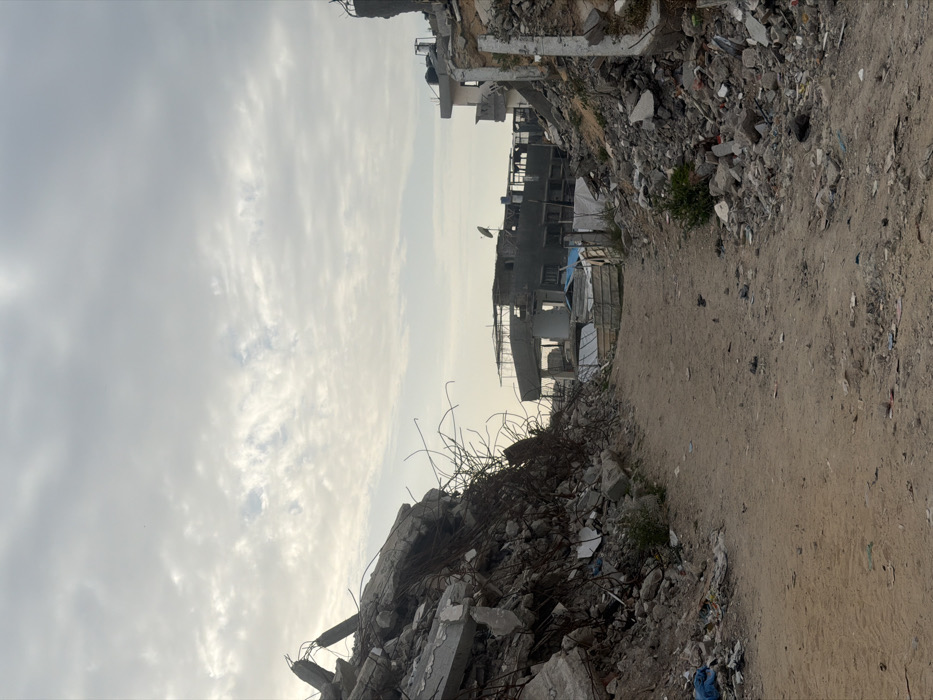 The image depicts a devastated urban landscape in Jabalia, Palestinian Territory, characterized by extensive structural damage and debris. A central, unpaved dirt path, approximately 2-3 meters wide, extends into the distance, narrowing slightly. On both sides of this path are large mounds of rubble, composed of broken concrete, twisted rebar, masonry fragments, and other building materials, indicating collapsed structures.
To the left, a substantial pile of debris forms a steep slope, showing exposed rebar protruding upwards and outwards. To the right, similar rubble piles line the path, with some standing, fractured wall sections visible further back. Small patches of sparse vegetation are discernible among the debris.
In the mid-distance, several buildings appear heavily damaged or partially destroyed. One building on the left horizon features an exposed framework, while another structure further along the path shows missing sections and exposed interiors. A dark, possibly partially collapsed, multi-story building is visible in the center-right background, with a small satellite dish mounted on its upper level. Minimal, makeshift structures, possibly constructed from corrugated metal, are visible near the distant buildings. The sky above is overcast with scattered clouds, and no individuals or active interactions are present in the scene.