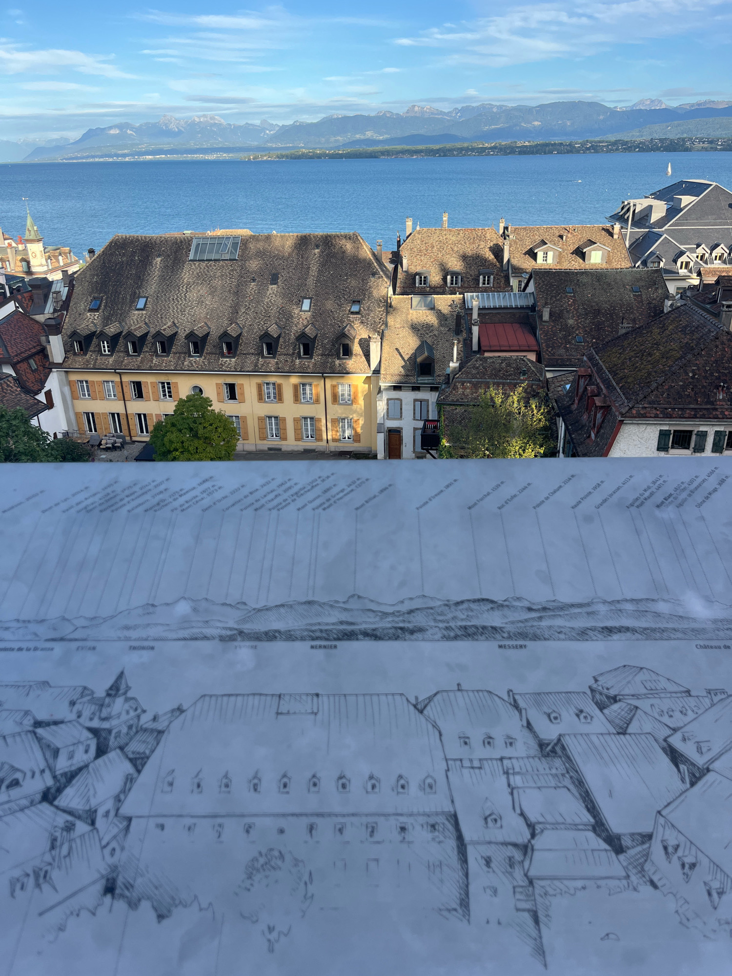 This image captures a panoramic view of Nyon, Switzerland, overlooking Lake Geneva and the distant French Alps. In the foreground, a large, detailed sketch is laid out, depicting a cityscape with numerous buildings and a detailed mountain range outline. Text annotations are visible on the sketch, labeling various peaks and locations, including "Evian," "Thonon," "Yvoire," and "Nernier." The actual cityscape of Nyon is visible in the upper portion of the image, characterized by terracotta-tiled roofs, mansard roofs, dormer windows, and multi-story buildings. A prominent yellow building with numerous windows and shutters stands out. Beyond the buildings, the vast expanse of Lake Geneva reflects the blue sky, and beyond that, a mountain range with snow-capped peaks forms the horizon under a clear blue sky with scattered clouds. The overall scene evokes a sense of historical and geographical context, with the sketch serving as an interpretive overlay of the real-world vista. There are no people or animals visible in the image.