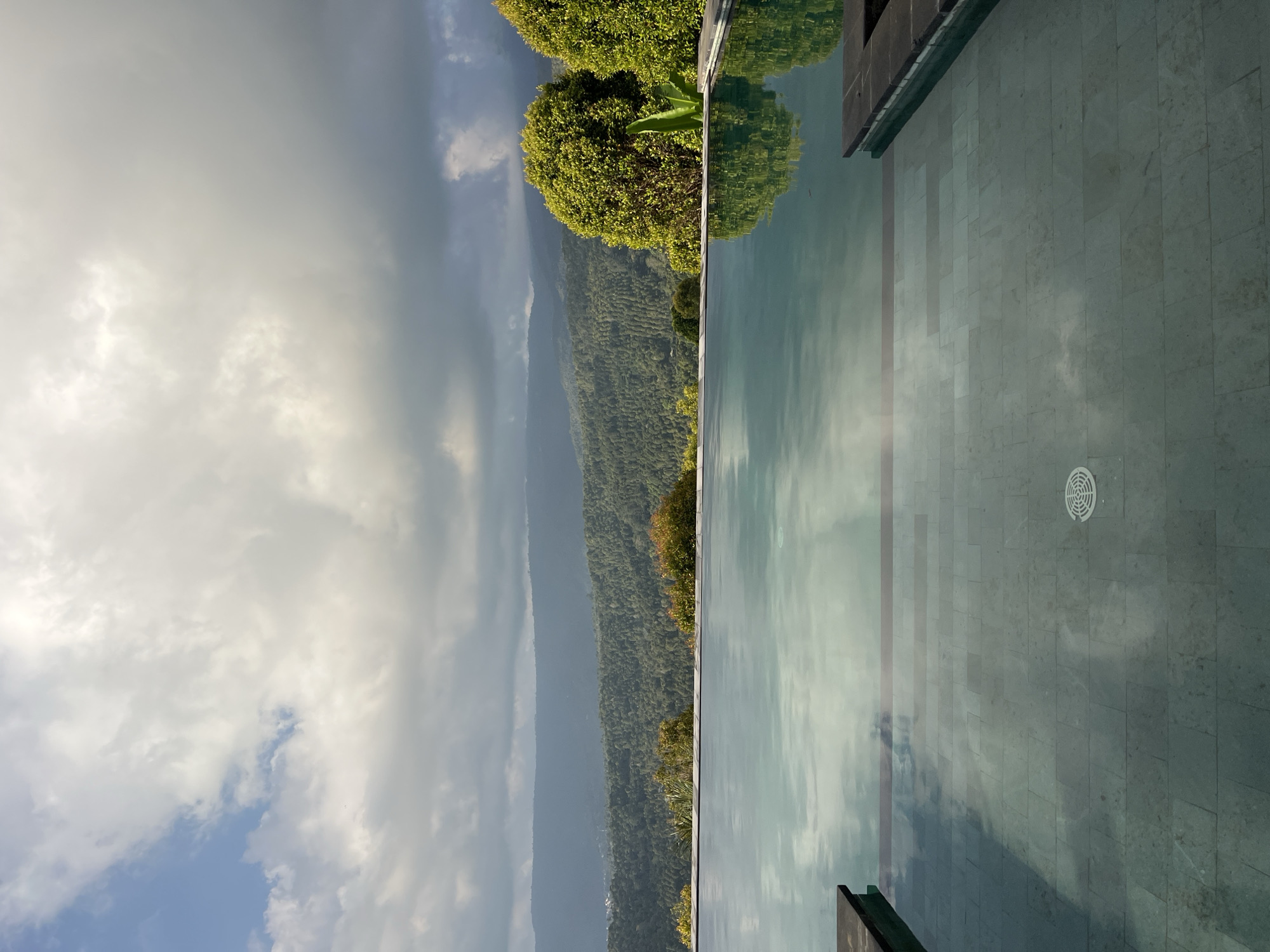 An infinity pool overlooks a lush, mountainous landscape in Busungbiu, Indonesia. The pool's surface reflects the sky, which is filled with dramatic, dark clouds and patches of blue. Beyond the pool, rolling hills covered in dense green foliage stretch into the distance, with a hazy mountain range forming the horizon. Two rounded, vibrant green trees stand prominently on the right side of the frame, their leaves catching the light. The pool is constructed with dark stone coping and a tiled bottom, visible through the clear water. A circular drain grate is visible on the pool floor. The scene appears calm and serene, capturing the natural beauty of the Indonesian countryside.