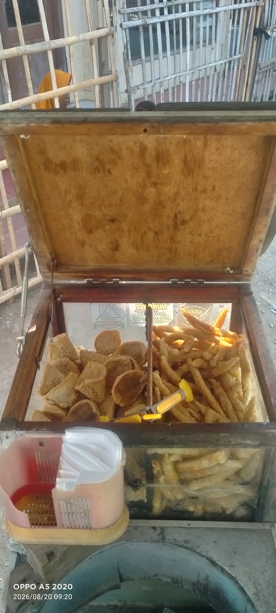 In the city of Sowan Kidul, Indonesia, a street food vendor's cart is displayed. The cart is made of wood and has a hinged lid. Inside, two compartments hold fried snacks. The left compartment contains a pile of irregularly shaped, golden-brown fried items, possibly tofu or fish cakes. The right compartment holds a larger quantity of slender, elongated, golden-brown fried items, resembling french fries or fried dough sticks. A yellow and silver tong rests on top of the fried items in the right compartment. In front of the cart, a pink and white plastic container, possibly for napkins or utensils, is visible. In the background, a person wearing a yellow headscarf is partially visible behind a bamboo fence. The ground appears to be a rough, grey surface. The lighting suggests it is daytime.