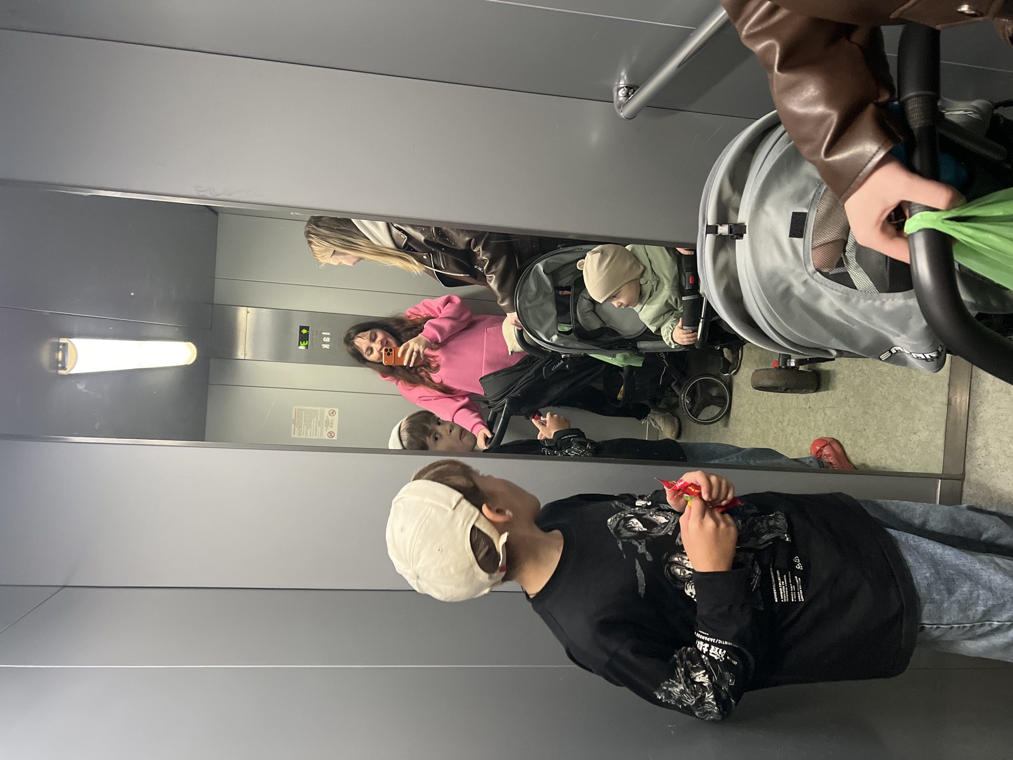 The scene depicts the interior of an elevator car, located in the city of Chaikovsky, Russia. The elevator features grey paneled walls and a large, reflective mirror surface on the right side. Overhead, a single long, cylindrical fluorescent light fixture illuminates the space.
In the foreground, a young boy with light brown hair, wearing a light-colored baseball cap reversed, stands with his back partially to the camera. He wears a black long-sleeved shirt with graphic prints that include Japanese text and stylized imagery, and light blue denim jeans. He holds a small red and yellow packaged snack item in his hands and is looking towards the right, into the mirror.
The mirror reflects a group of people and objects:
- A woman with long dark hair, wearing a pink hooded sweatshirt, is holding a smartphone with an orange case up, appearing to take a mirror selfie. She is smiling.
- To her right stands another woman with long blonde hair, wearing a brown leather-look jacket over a light-colored hoodie. Her right arm extends to hold the handle of a grey child's stroller. A green plastic shopping bag is hanging from the stroller handle.
- Seated in this stroller is a baby or young toddler, wearing a light green jacket and a beige beanie hat, looking towards the right in the reflection.
- The reflection of the foreground boy is visible to the left of the woman in pink, showing his face looking towards the group.
- Environmental details visible in the mirror include a metallic elevator control panel with a glowing green "E" and an upward arrow, alongside reversed Cyrillic text (likely a floor indicator), and a small white warning sign with red text and symbols indicating 'no smoking' and 'no food/drink'.
A metallic handrail is partially visible on the far right wall, near the bottom of the frame. A second grey child's stroller is partially visible in the lower right foreground, obscuring its occupant. The group appears to be waiting or traveling within the elevator.