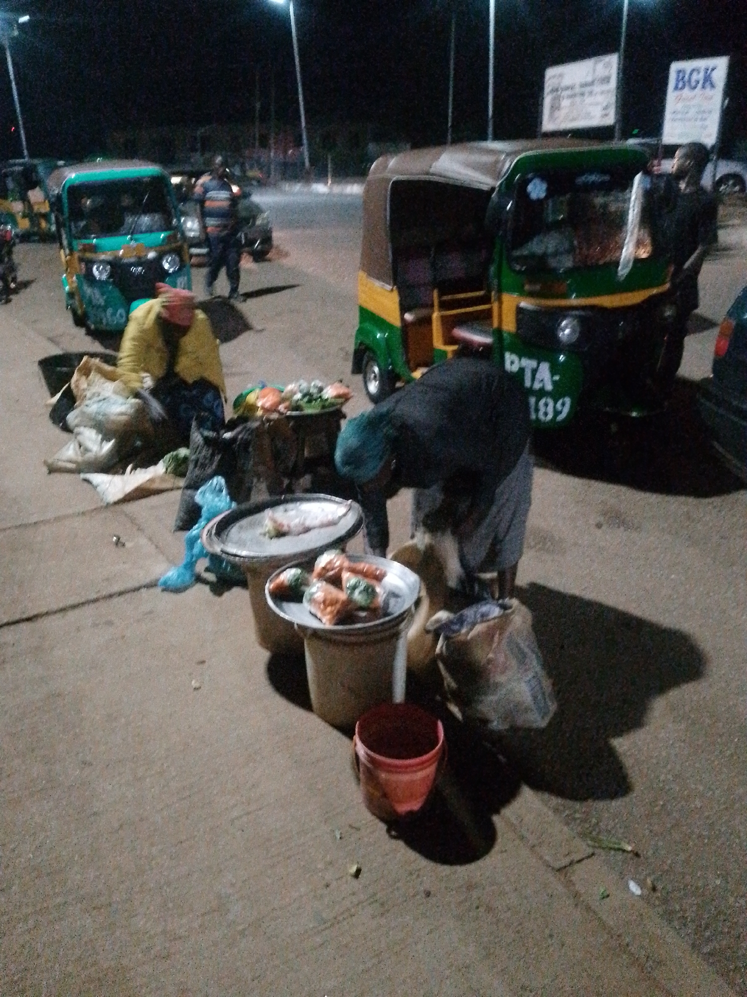This is a nighttime photograph taken in the city of Bukuru, Nigeria. The scene depicts a street market with several auto-rickshaws, commonly known as Keke Napep. In the foreground, a person dressed in a dark blue shirt and grey pants is bent over a collection of produce, which appears to be vegetables and fruits, displayed on trays and bowls. There are several large sacks and bags of goods scattered around the vendor.
To the left, another auto-rickshaw, painted green and yellow, is visible. A person in a yellow garment and a red head covering is seen partially obscured behind it, next to sacks of produce.
In the background, more auto-rickshaws are parked or in motion, and streetlights illuminate the area. Signs are visible on poles in the distance, one of which displays "BGK" in large letters. The ground is paved, and the overall impression is of a bustling, albeit dimly lit, nighttime marketplace.