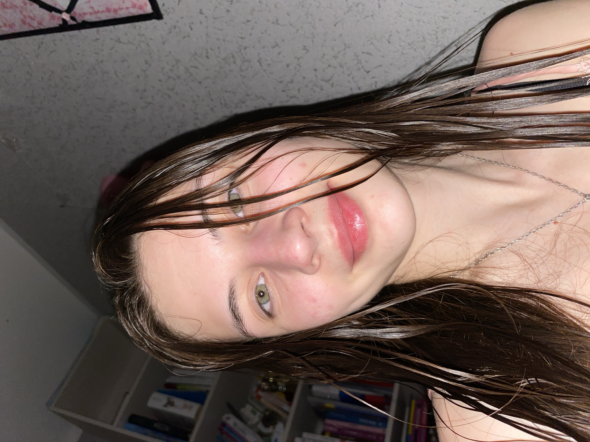 A close-up portrait, originally captured with a rotated perspective, shows a young individual indoors in Votkinsk, Russia. The subject has fair skin, dark brown hair, and light green eyes. Their wet, dark brown hair falls across their forehead and shoulders. They are looking directly at the camera with a neutral expression. Their lips are full and have a glossy pink tint, possibly from lip gloss or lipstick. Minor redness is visible on their nose and cheeks. A delicate silver-colored chain necklace adorns their neck, and a black strap, likely from clothing, is visible on the upper right side of the frame. The background is softly out of focus, revealing a textured wall surface on the left and a shelving unit filled with books and various objects on the right. A dark object with pink elements, possibly a poster or decoration, is partially visible in the top left corner. The lighting suggests an indoor setting, possibly at night or in dim conditions. No visible text is present in the image.