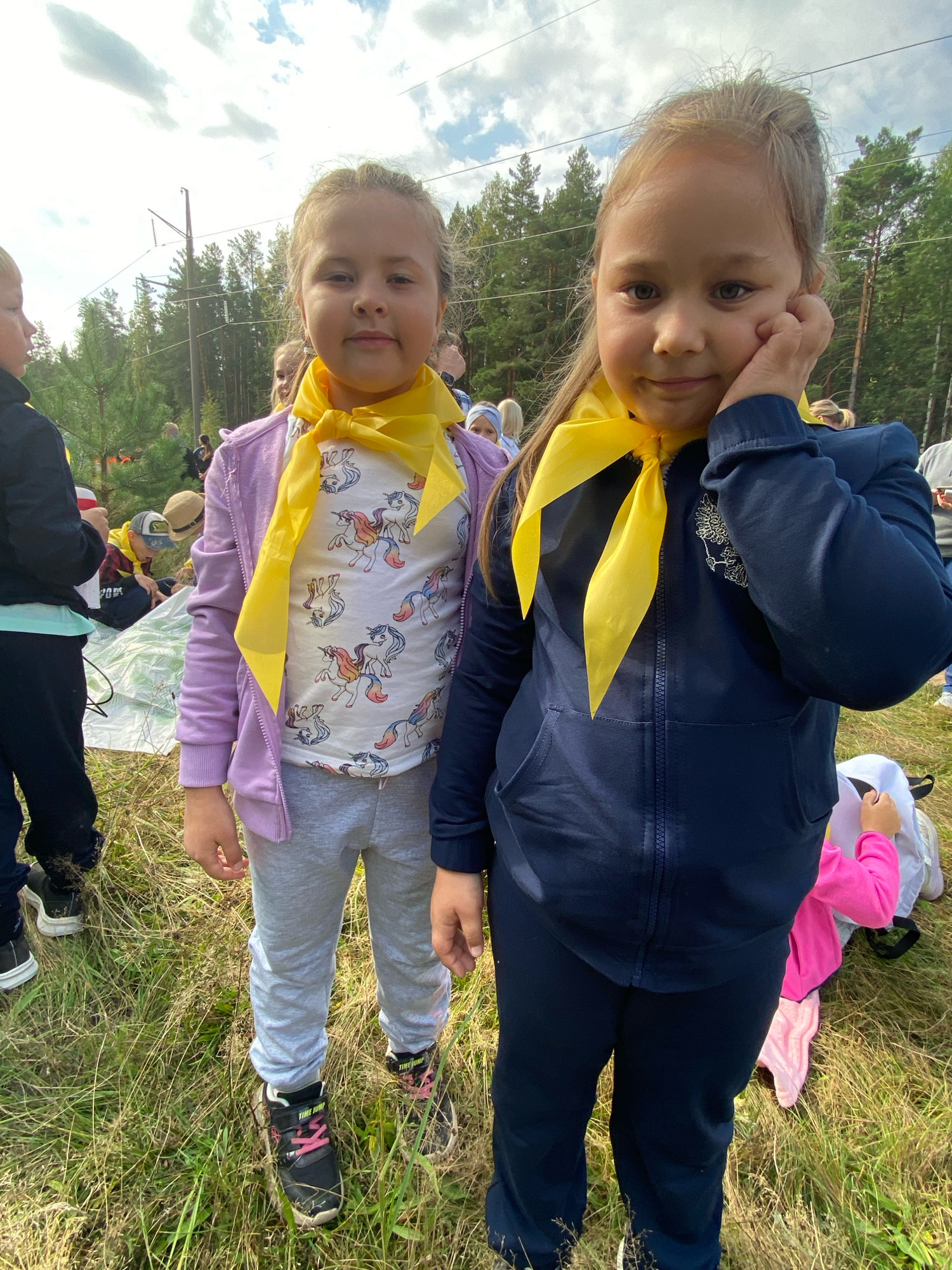 The image shows two young girls wearing yellow scarves in an outdoor setting, likely in Noviy, Russia. The girl on the left wears a purple jacket over a white t-shirt with a unicorn print and grey sweatpants. The girl on the right wears a dark blue tracksuit. Both girls have light brown hair and are looking towards the camera. In the background, there are trees and a cloudy sky. Other children and adults are visible in the background, some sitting on a mat. The environment appears to be grassy and somewhat wooded.