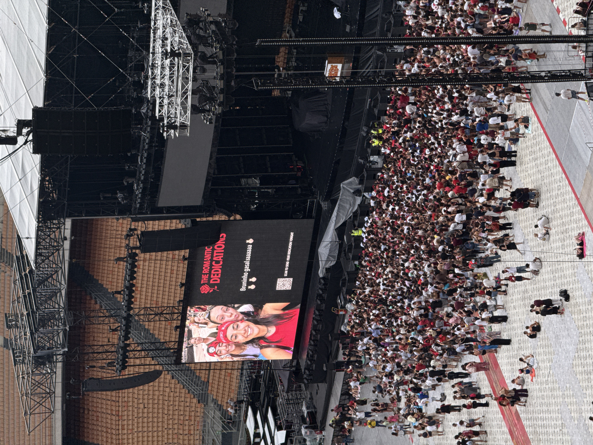 An aerial, wide-angle shot shows a large crowd of people gathered in a stadium in Milan, Italy, for a concert. The stadium has tiered seating in the background, largely empty but with a few individuals visible. In the foreground, a dense crowd of concertgoers is milling about on a paved area. The crowd is diverse in age and attire, with many wearing red clothing, possibly indicating team or fan affiliation. Some people are sitting on the ground, while others are standing and talking.
Dominating the scene is a large stage setup with a massive screen displaying a photograph of four young women smiling and taking a selfie. Text on the screen reads "THE ROMANTIC DEDICATION" with a rose icon, followed by "Bruninho gasalssssaa" and a QR code with the instruction "SCAN TO SEND YOUR DEDICATION TONIGHT". The stage itself is complex, with multiple levels, lighting rigs, and sound equipment. A black tarp covers a central area of the stage.
The overall atmosphere appears to be festive and anticipatory, with the crowd waiting for the event to begin. The lighting suggests it is daytime, though the stadium structure casts shadows on parts of the crowd. The image captures a moment before a large-scale public event, highlighting the scale of the gathering and the visual spectacle of the concert setup.