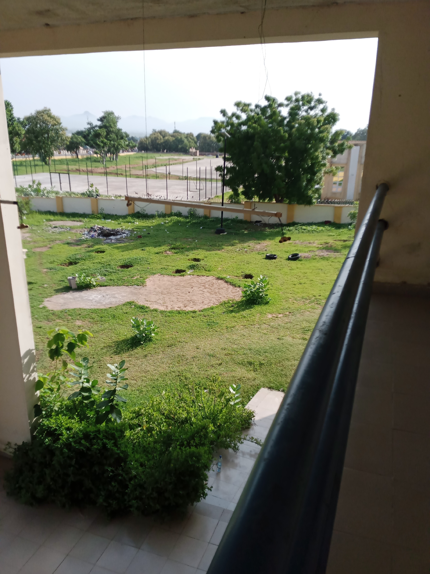 The image depicts an outdoor scene, likely a courtyard or grounds, viewed from a covered walkway. In the foreground, lush green bushes and plants are visible, with some lighter-colored leaves on taller plants. The ground is covered in green grass with several circular patches of bare soil or sand. A pile of debris, including what appears to be plastic and other waste, is present in the middle ground. Beyond the grassy area, a fence with vertical posts and horizontal wires separates the grounds from a paved area or road. In the background, several trees with dense green foliage are scattered across a landscape that includes distant hills under a hazy sky. The architecture of the building from which the photo is taken is visible on the right, with a dark metal railing running along the walkway. There are no people visible in the image. The scene suggests an undeveloped or slightly neglected outdoor space.