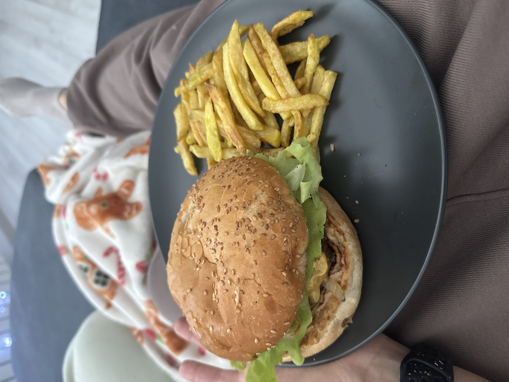 The image shows a grey plate holding a burger with lettuce and a pile of french fries. The burger bun is sesame-seeded. A person is holding the plate, their wrist visible with a black smartwatch. They are wearing a brown top. In the background, part of a blanket with a cartoon pattern is visible, along with a glimpse of a leg with a white sock. The location cannot be determined from the image alone. The city of Sofia, Bulgaria, cannot be confirmed.