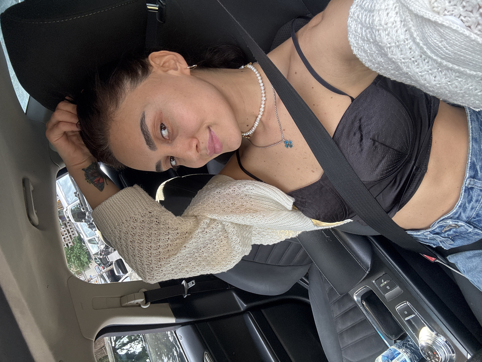 A woman sits in the driver's seat of a car, with her right arm raised and hand on her head. She is wearing a black camisole, blue denim jeans, and a white knitted cardigan draped over her shoulders. She has a pearl necklace with a blue charm. The car's interior, including the black leather seats, dashboard, and seatbelt, is visible. Outside the car, through the window, parked cars and buildings can be seen, indicating an urban setting.