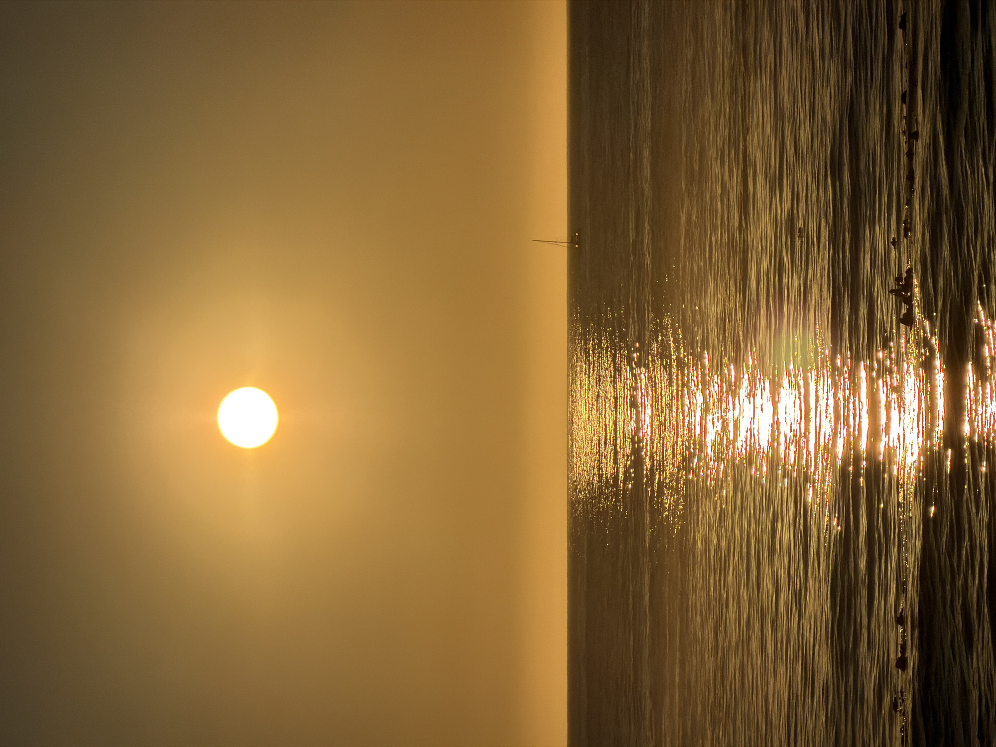 The image depicts a serene ocean sunset over the waters of Cape Town, South Africa. The sun, a bright orb, is positioned in the upper center of the frame, casting a warm, golden glow across the sky and water. The sea is calm with gentle waves, and a prominent reflection of the sun stretches across the surface, creating a shimmering path. In the distance, a solitary sailboat with its mast visible glides across the water, adding a sense of tranquility to the scene. The horizon line is clearly defined between the sky and the ocean. Some dark, indistinct shapes, possibly rocks or debris, are present in the foreground, breaking the smooth expanse of the water. The overall atmosphere is peaceful and highlights the natural beauty of a sunset over the ocean.