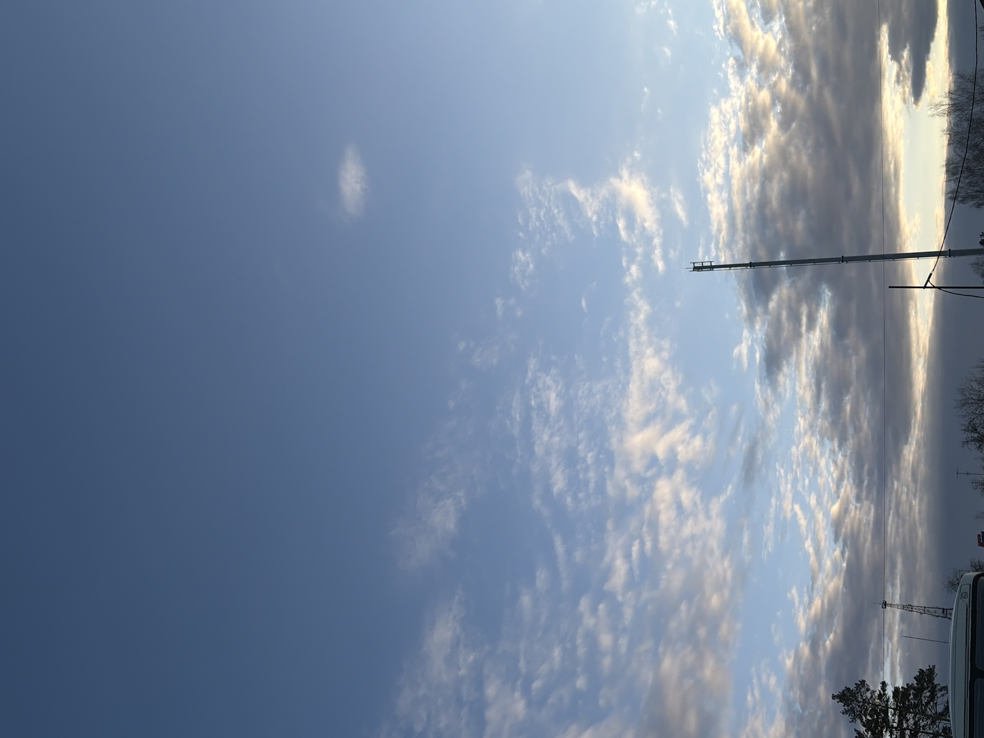 The image is a vertically oriented outdoor shot capturing a partially cloudy sky over a location in the city of Chaikovsky, Russia. The sky transitions from a clear, deep blue on the left to a lighter blue with scattered altocumulus clouds on the right. A faint, circular cloud is visible in the upper-left. A thin, horizontal structure, possibly a crane or utility pole, extends across the upper-right portion of the frame, partially obscured by the clouds. The edges of trees are discernible along the right edge of the frame, and a glimpse of a vehicle or structure is visible in the lower-right corner. Electrical wires run across the scene to a communications tower.