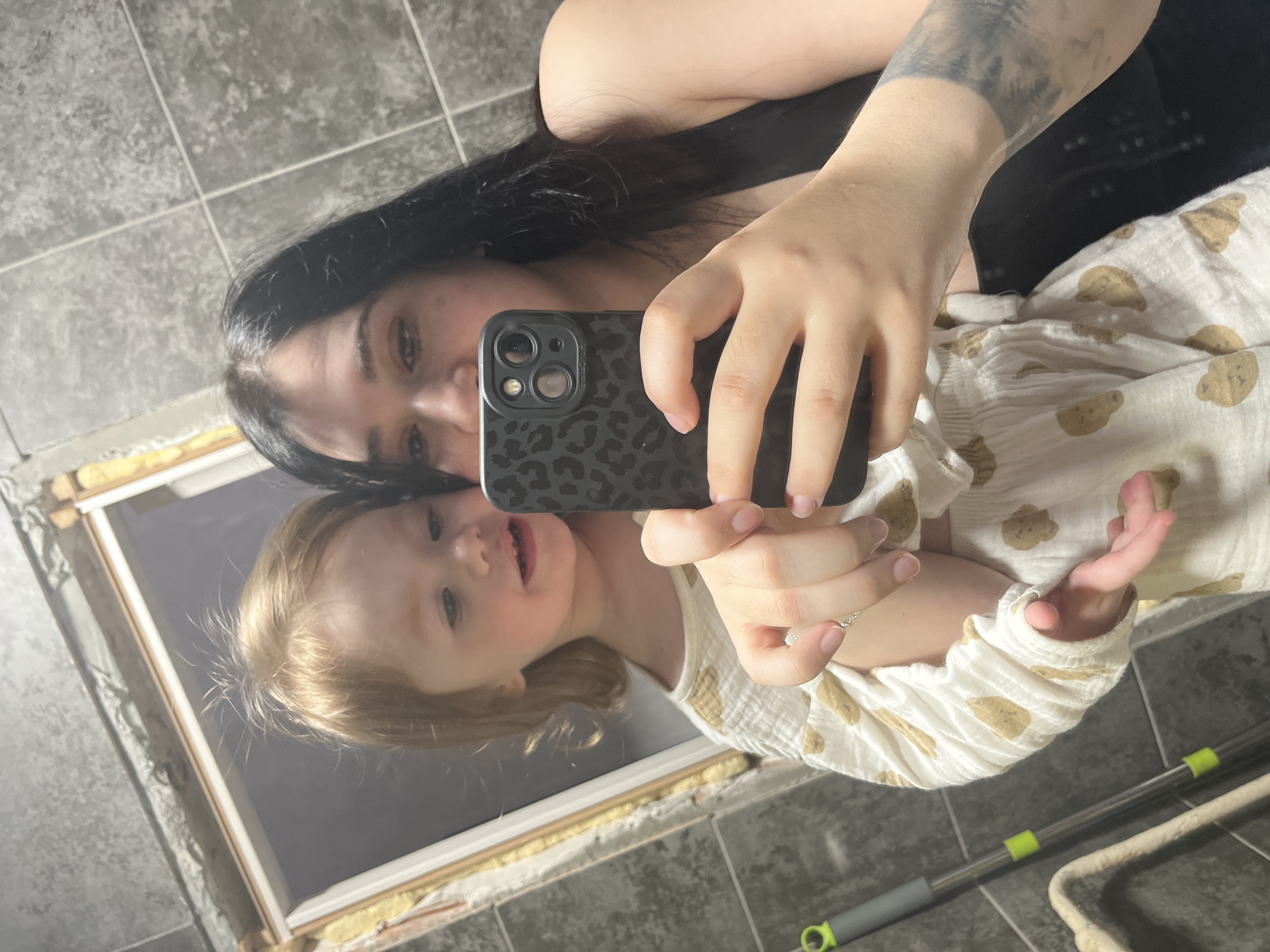An adult female with long dark hair and a tattoo on her left forearm is depicted taking a mirror selfie with a black smartphone featuring a leopard-print case. She wears a black tank top. She is holding a young child with light, reddish-blonde hair, dressed in a white sleepsuit patterned with brown bear motifs. The child's gaze is directed to the left, with a slight smile. The background features dark grey, square tiled walls and an unfinished window opening with a white frame, exposed yellow insulation, and visible concrete. A grey mop with green accents leans against the wall in the lower-left foreground. The scene is an indoor environment in Chaikovsky, Russia, consistent with a utility space or room under renovation.