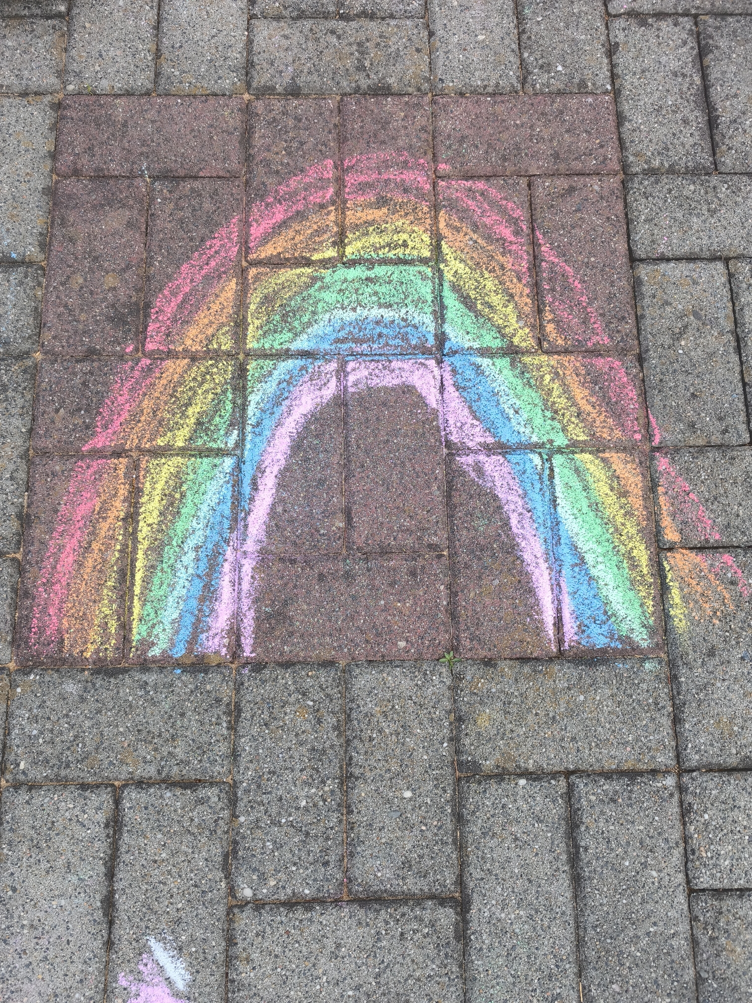 The image captures a vibrant chalk rainbow drawn on a paved outdoor surface. The rainbow arches across several rectangular paving stones, predominantly reddish-brown in color, with its ends extending onto surrounding gray stones.
The rainbow itself features a sequence of colors, appearing from outermost to innermost as pink, orange, yellow, green, light blue, and a lighter purple or pink for the innermost arc. The chalk marks show the rough, granular texture of the paving stones, indicating a direct application to the concrete or stone surface. The lines of the rainbow are broad and hand-drawn, giving it a playful, informal appearance.
The paving stones are laid in a grid pattern, with visible grout lines between them. The surrounding stones at the top, bottom, and sides of the frame are gray, contrasting with the central reddish-brown area where the bulk of the rainbow is situated.
The scene is brightly lit, suggesting it is daytime, and the colors of the chalk are vivid, although slightly diffused by the textured surface. There are no people or other identifiable objects in the frame, and no visible text is present. The image conveys a sense of simple, creative outdoor activity.