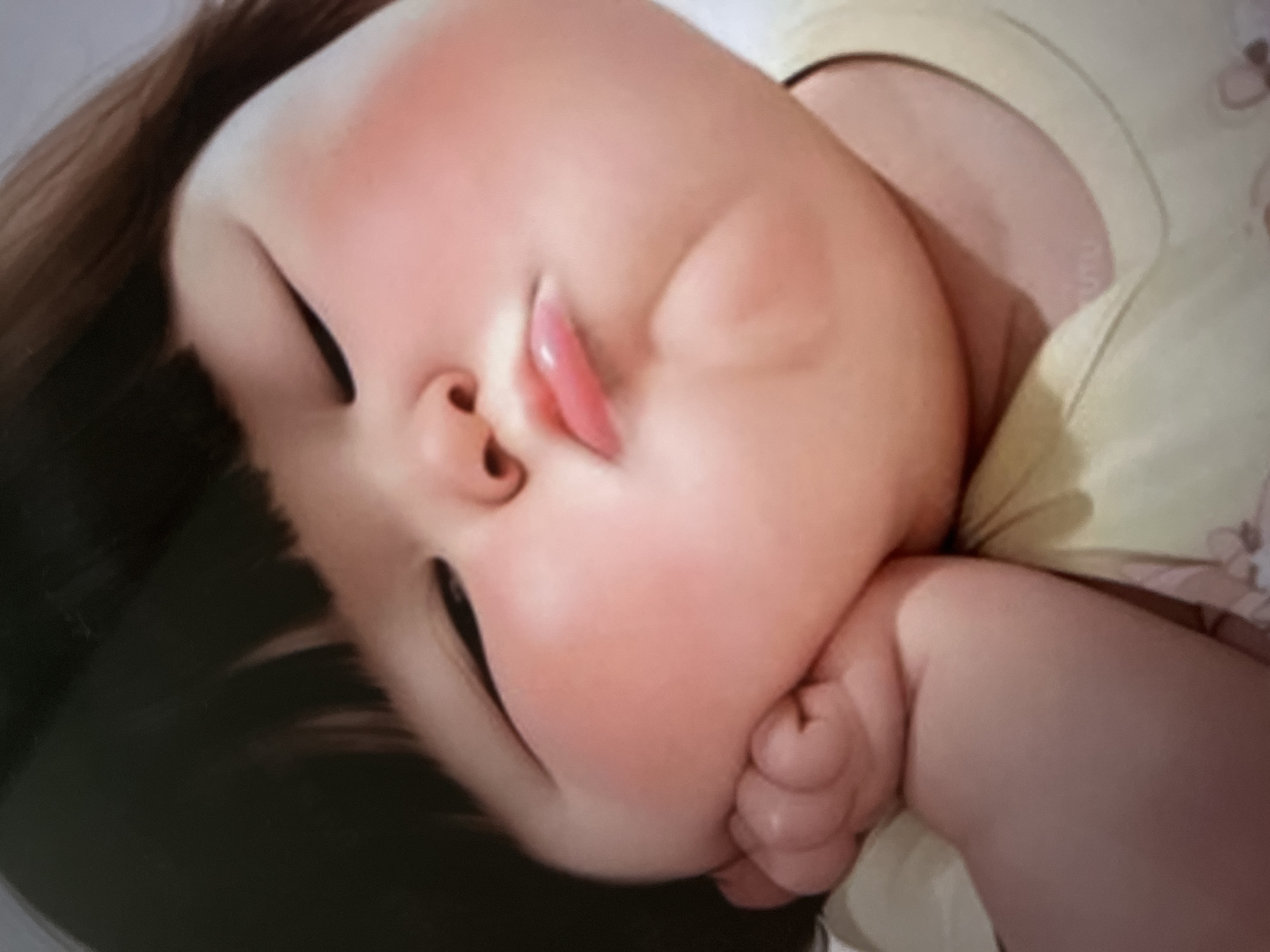 A close-up, eye-level, full-frame shot shows a digital illustration of a plump baby girl with her eyes closed, resting her head on her hand. She has dark hair styled in a bob, with bangs covering her forehead. Her cheeks are flushed pink, and her lips are pursed. The background is blurred and appears to be a light-colored surface with a hint of fabric pattern. The image is rendered with soft lighting, creating a gentle and tender atmosphere. The baby is wearing a light yellow t-shirt.