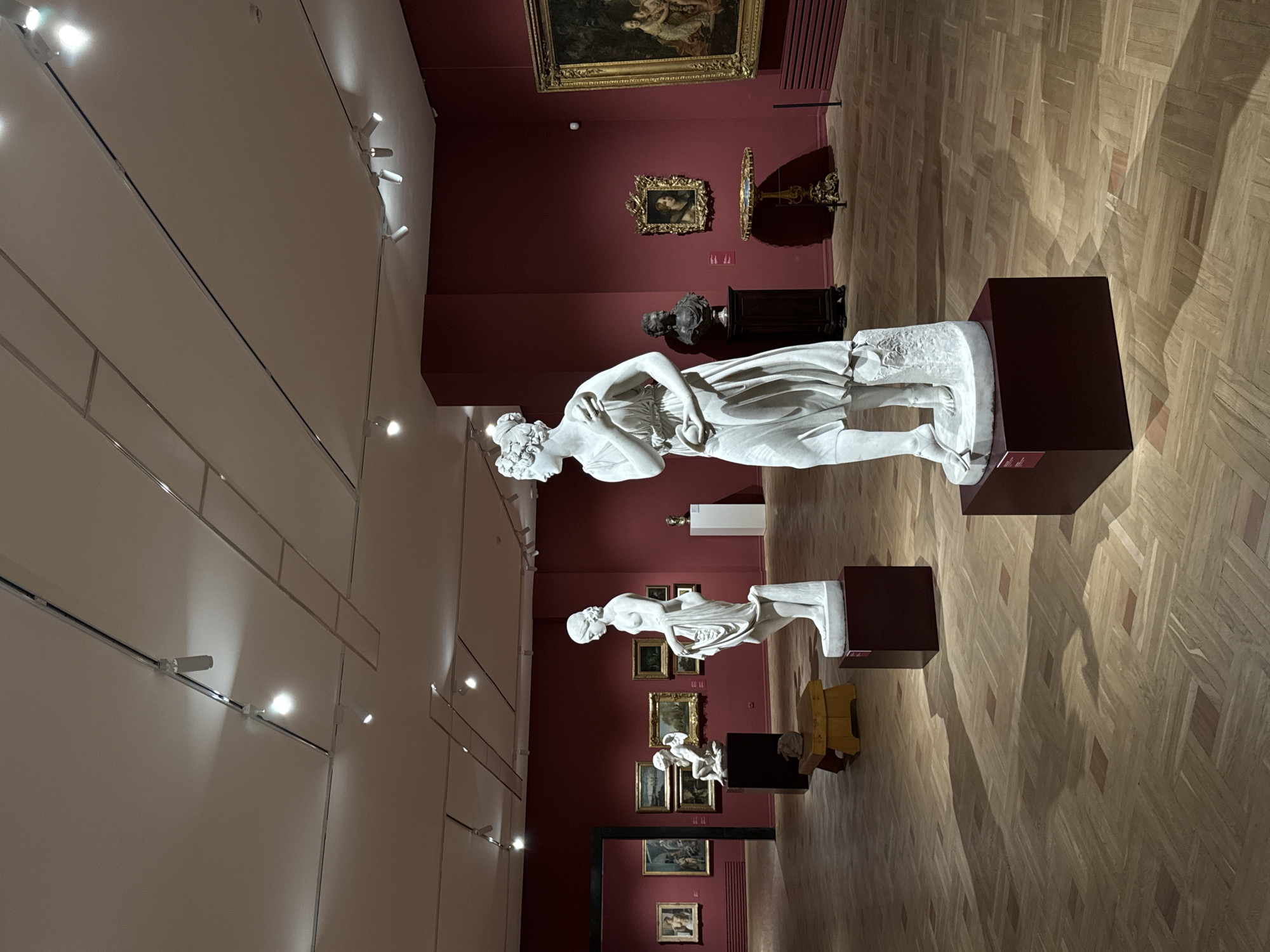 The image shows the interior of a museum gallery with burgundy walls and a polished wooden floor. Several white marble statues are displayed on dark rectangular pedestals. In the foreground, a statue of a female figure stands draped in cloth, holding something in her hands. Further back, another female statue is visible. Other sculptures and framed paintings are displayed on the walls and in other parts of the gallery. Track lighting illuminates the space from the ceiling. The scene suggests an art exhibition or a permanent collection display. This scene is located in the National Gallery of Armenia in Yerevan, Armenia.