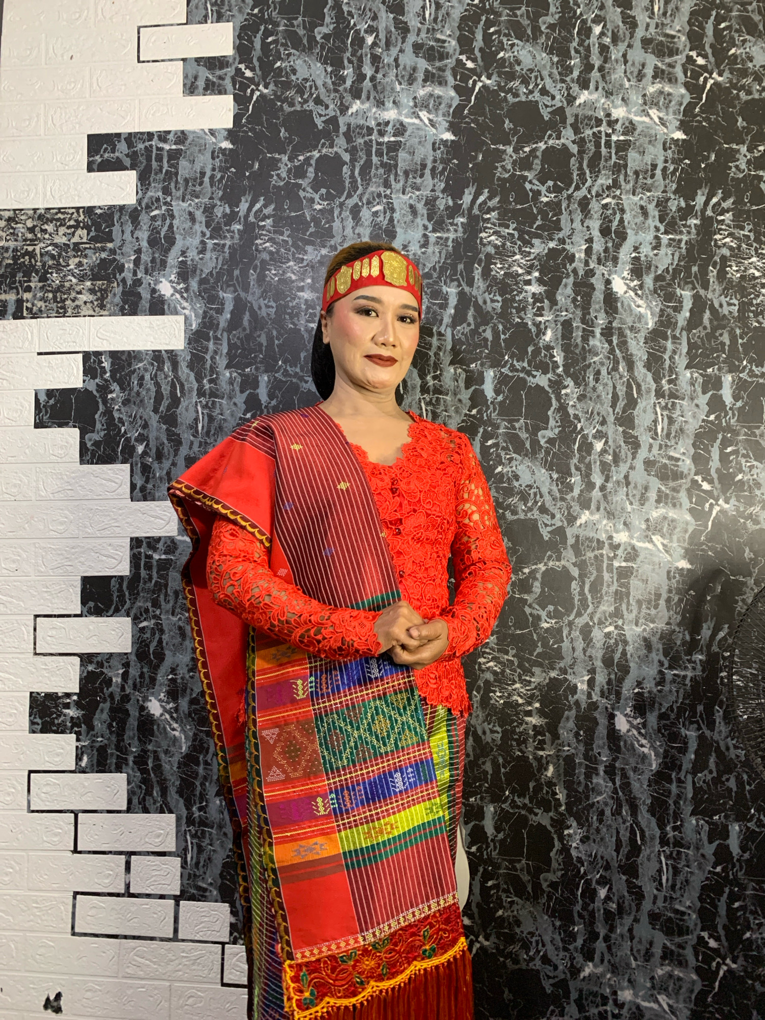 A woman stands in front of a wall with a black marble pattern and a white brick pattern. She is wearing a red lace top with intricate embroidery and a red and colorful striped sash draped over her shoulder and around her waist. She also has a red and gold headband on her head. Her hands are clasped in front of her. The background is dark, with the marble pattern creating a dramatic effect. The brick pattern on the left side of the image provides a contrasting texture. It is not possible to determine the location within Malang, Indonesia from the image.