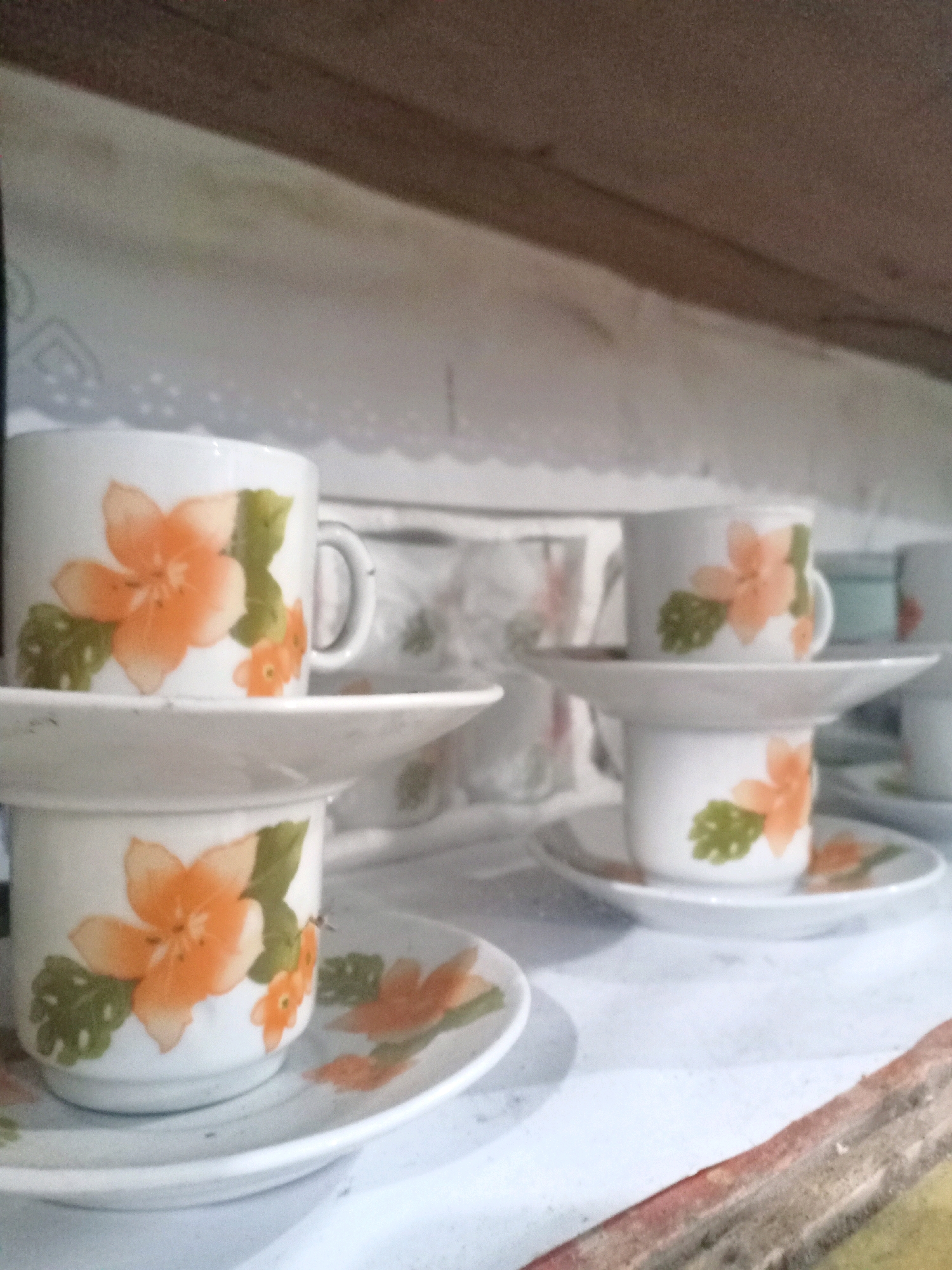 The image shows a close-up view of a shelf displaying several ceramic teacups and saucers with a floral pattern. The teacups are white with orange flowers and green leaves painted on them. There are multiple teacups and saucers stacked and arranged on the shelf, suggesting they are part of a set. In the foreground, a teacup and saucer sit prominently on a white surface, possibly a tablecloth or the shelf itself. The background is slightly blurred, but the overall scene appears to be an indoor setting, likely a kitchen or dining area, given the presence of teaware. The lighting suggests it is daytime, and there are no indications of specific weather conditions. The arrangement of the teacups is neat and orderly. There is no visible text in the image. The context provided is Jarit, Indonesia, which might indicate the origin or style of the teaware, though this is not explicitly depicted in the visual content.