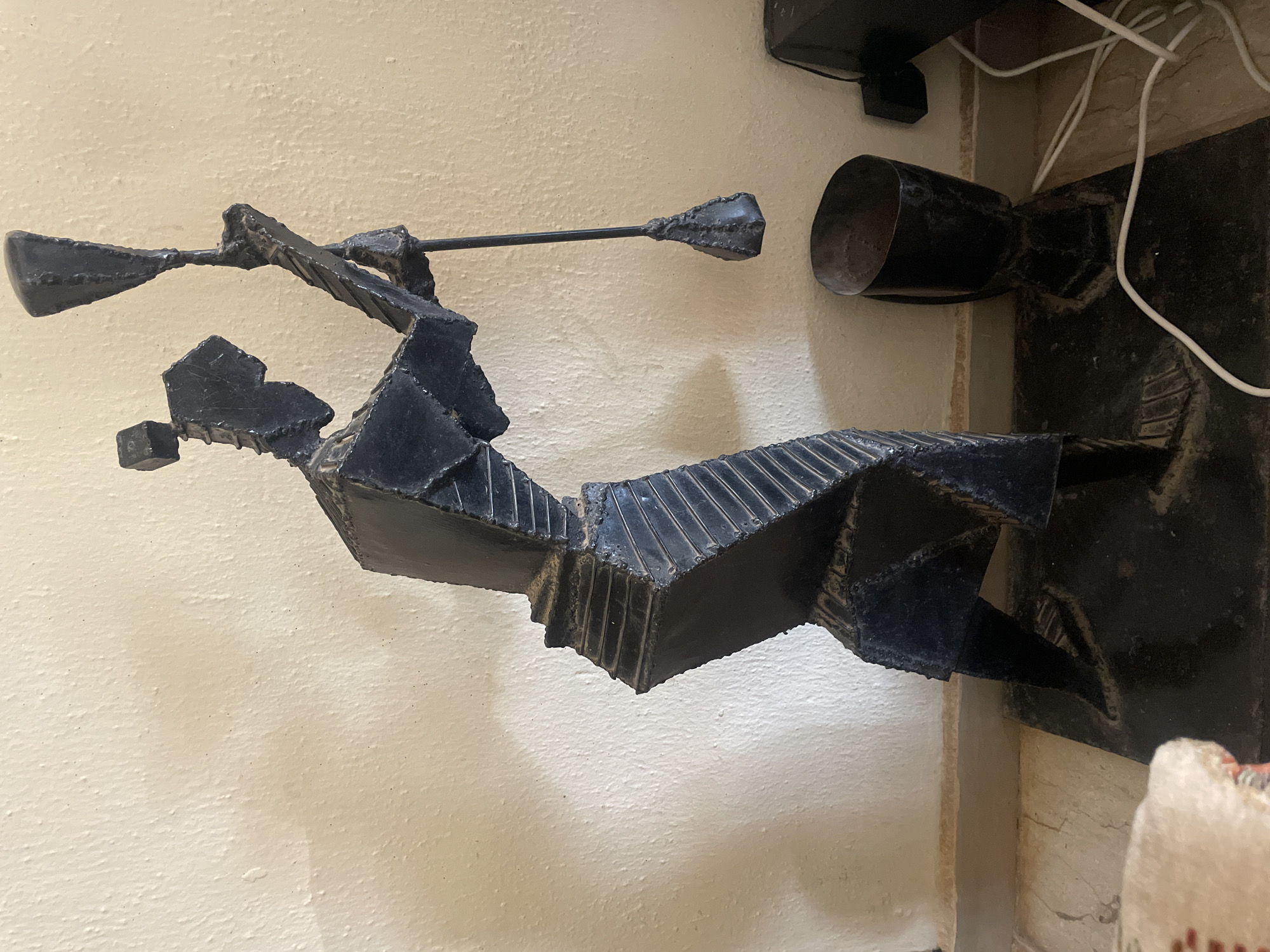 The image shows a welded metal sculpture of a knight holding a lance, positioned against a light beige wall. A black cylindrical object sits on a dark surface with white cords. The location cannot be determined from the image.