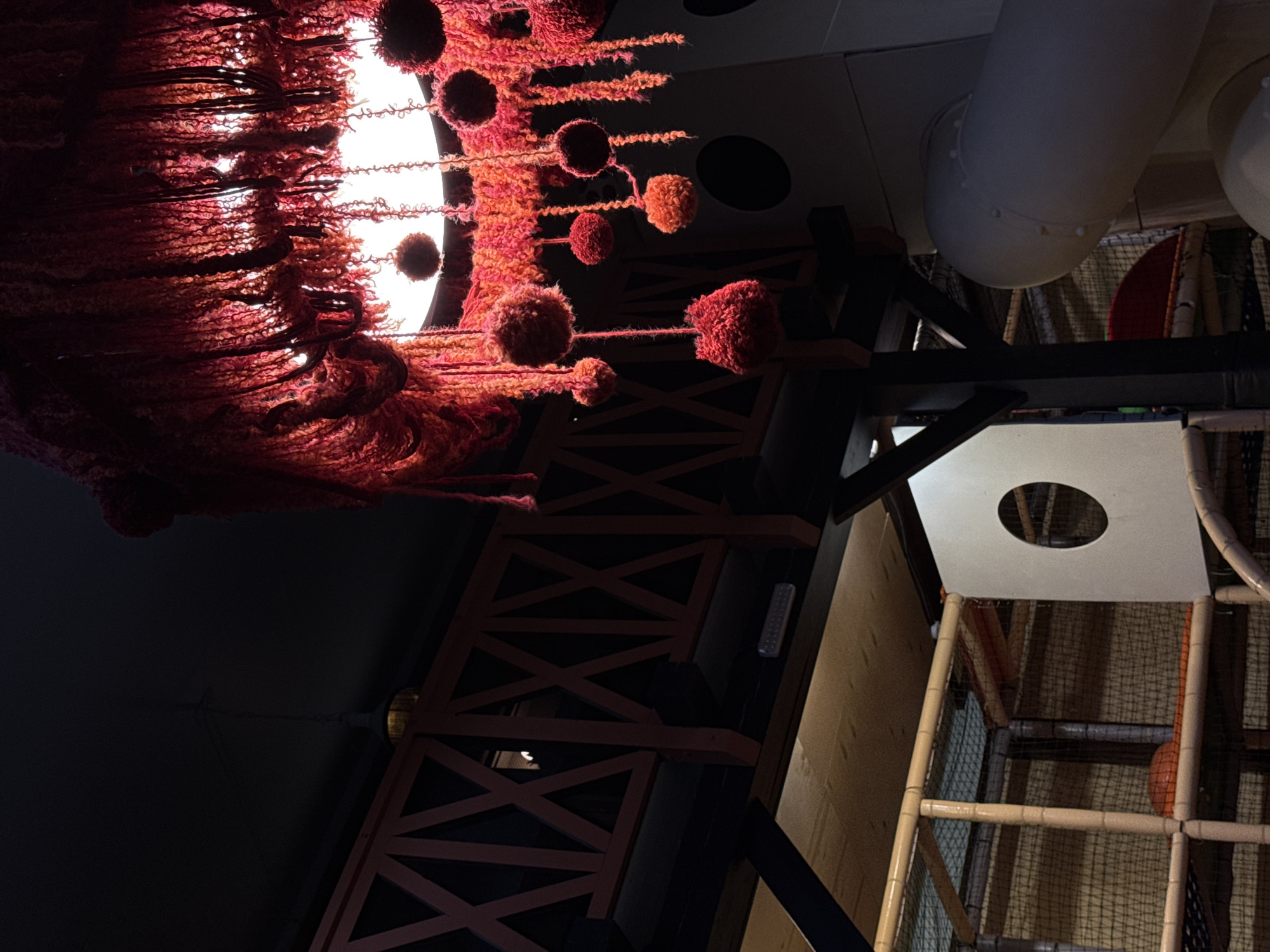 The image depicts an indoor playground structure, possibly in Izhevsk, Russia, but this cannot be confirmed from the image alone. A decorative light fixture dominates the upper left. It is composed of a circular array of light, obscured by dense, reddish-brown fibrous material and punctuated with spherical pom-pom-like decorations. To the right, the ceiling structure features a cylindrical duct and a black beam with decorative red X-bracing. Below, there is a white platform with a circular opening, part of the playground's structure. The supporting framework is a network of tan-colored tubing and netting. An orange ball is visible within the netting. The image is taken from a low angle, looking up into the structure.