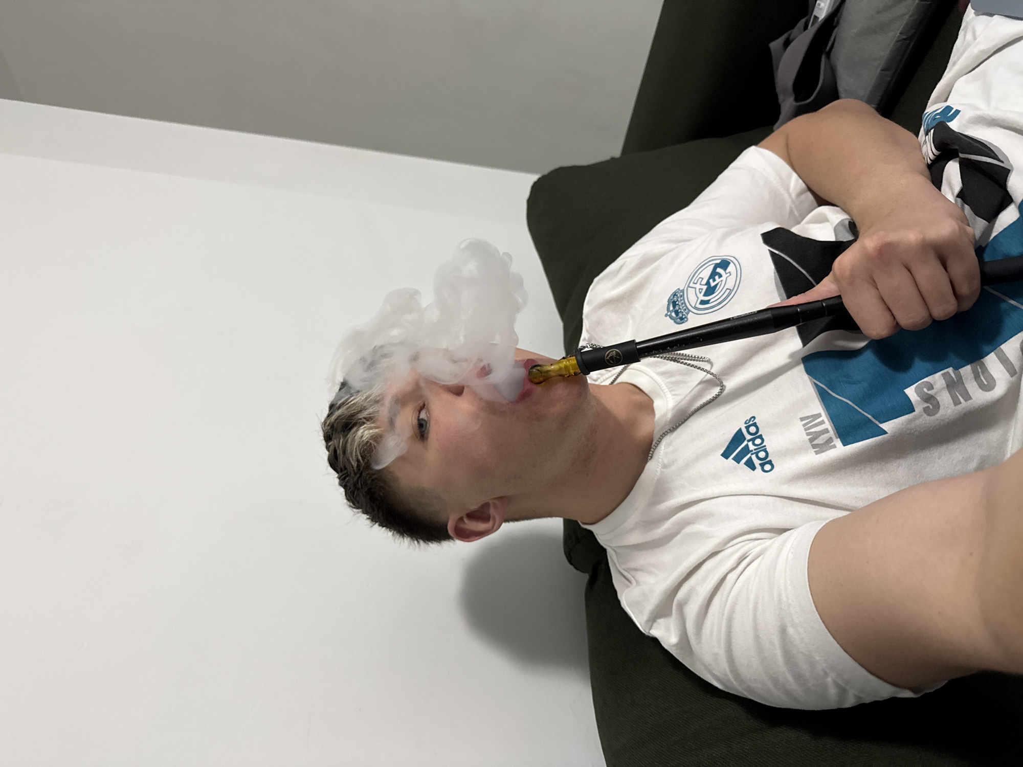 The scene is an indoor environment, likely a residential room. While the city is identified as Plovdiv, Bulgaria, the specific location within the city cannot be determined from the visual evidence.
A young adult male, with short dark hair featuring lighter bleached tips, occupies the central foreground. He is seated on a dark green upholstered sofa or armchair, facing slightly towards the camera. He wears a white short-sleeved t-shirt displaying a blue Adidas logo, the word "KYIV," and a Real Madrid crest. A silver-colored chain necklace is visible around his neck.
The male is actively exhaling a significant cloud of white smoke from his mouth, generated by a black hookah pipe. The hookah's mouthpiece, colored yellow and translucent, is held to his lips. His right hand grasps the black stem of the hookah pipe.
The background consists of a plain white wall on the left, meeting a lighter grey wall or surface at an obtuse angle on the right. A folded grey garment or cloth is partially visible on the sofa to the right of the subject.