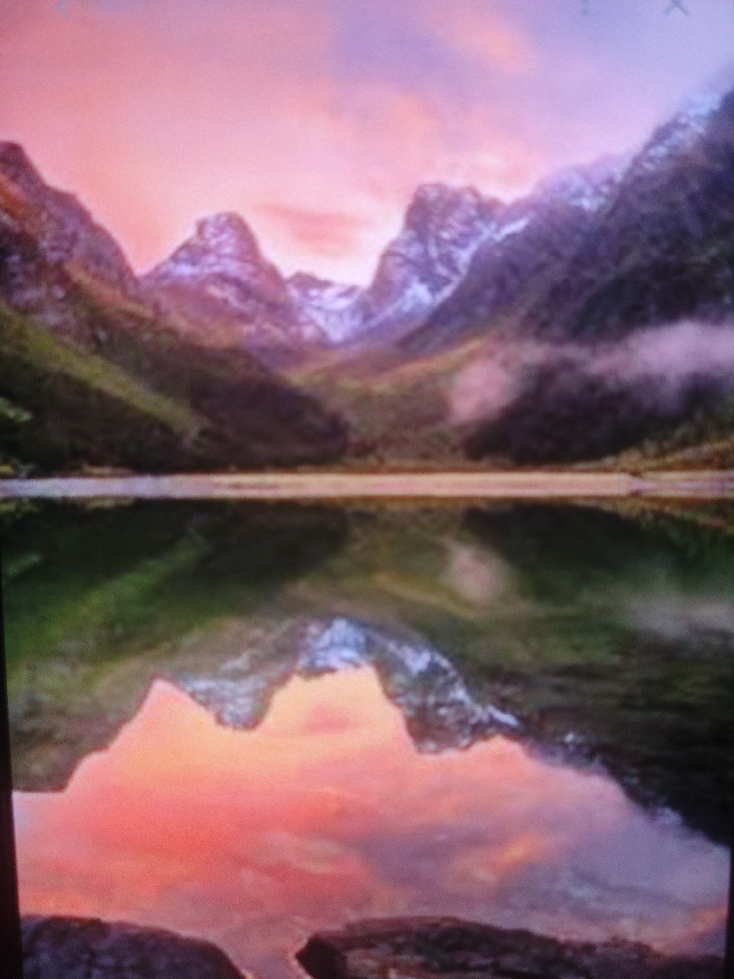 The image depicts a scenic mountain landscape with a lake reflecting the sky and mountains. The sky is painted with hues of pink and purple, suggesting either a sunrise or sunset. Snow-capped mountains rise in the background, their slopes partially covered in green vegetation. Mist or clouds drift around the mountain peaks. The lake in the foreground is calm, mirroring the dramatic colors of the sky and the imposing shapes of the mountains. The overall impression is one of natural beauty and tranquility.
This image does not depict the city of Armavir, Russia. The scene is a natural landscape, likely in a mountainous region, and there are no urban elements or structures visible that would associate it with Armavir.