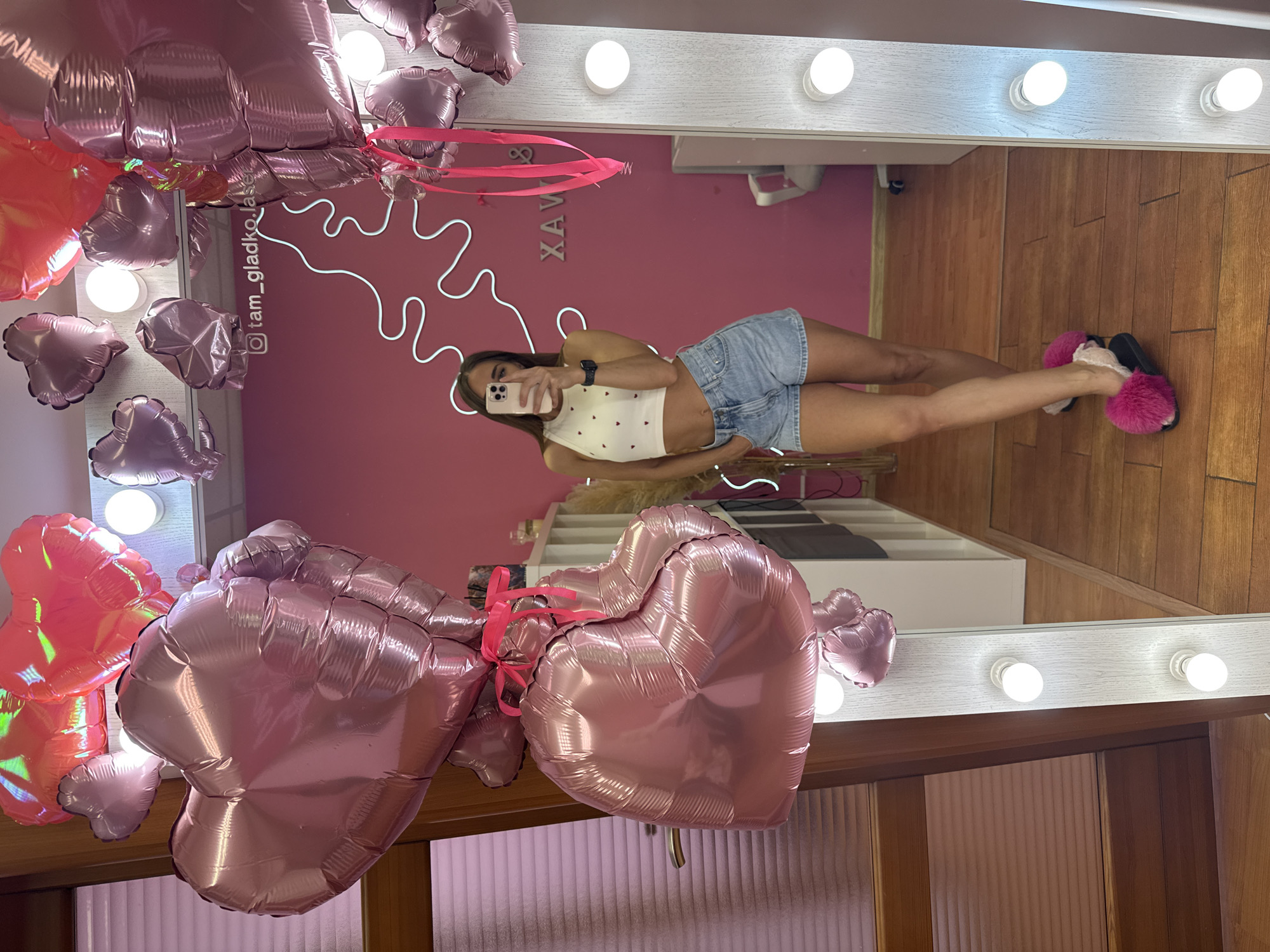 A woman is standing in front of a mirror, taking a selfie. She is wearing a white crop top with small red hearts and denim shorts. She has on pink fuzzy slippers. The mirror is illuminated by a ring of light bulbs. Behind her, there are several pink heart-shaped balloons. The room has pink walls and wooden floors. A neon sign on the wall reads "XAV & WAX". The watermark on the wall is "@tam_gladko.laser". This is a selfie taken in a beauty salon or studio.