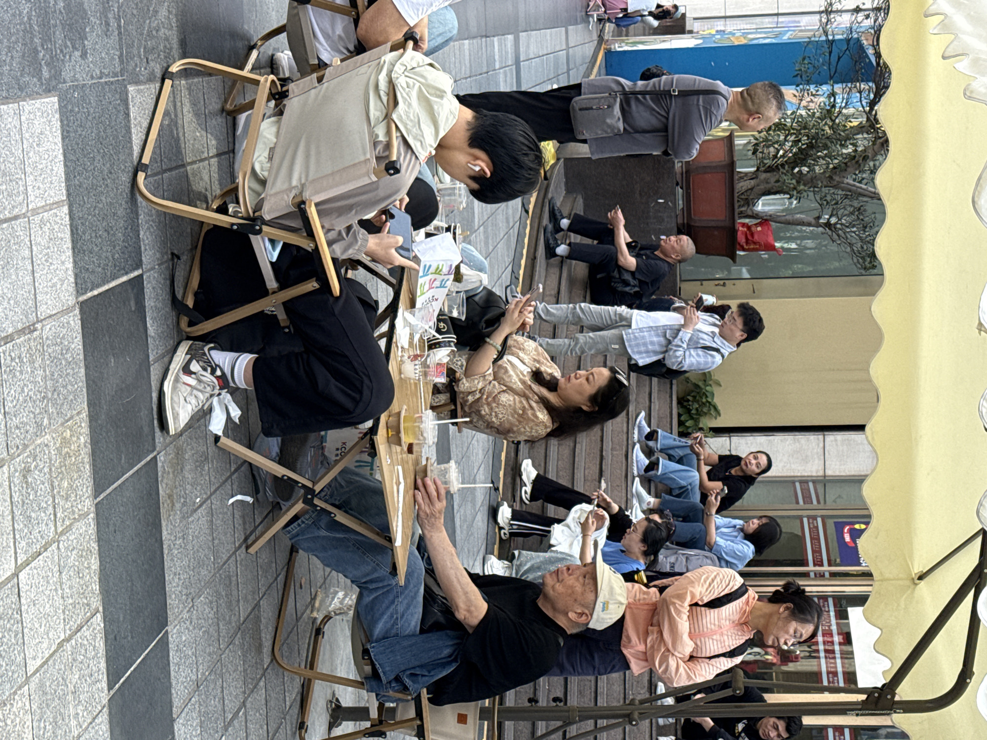 This full-shot, daytime photograph captures a vibrant street scene in an urban area, likely a pedestrian zone or sidewalk in China's Yuzhong District. Numerous people of various ages, from young adults to seniors, are gathered outdoors, predominantly seated and engaged in relaxed activities.
In the foreground, a group of individuals occupies portable camping-style chairs and small wooden tables. A young man on the left, wearing earbuds, is focused on his phone, while an older man in a light cap sits nearby, looking upwards. Several women are seated on low steps or the paved ground further to the right, many also absorbed in their mobile phones. A large yellow market umbrella provides shade to some of these individuals, indicating sunny weather.
Most people are dressed in casual attire, including t-shirts, jeans, and light jackets, and a common activity is the use of mobile phones. Beverages in plastic cups are visible on the tables, suggesting a casual outdoor cafe setting or simply a place for people to rest and socialize. The ground is paved with grey and black tiles. In the background, modern building facades with windows and some indistinct signs in Chinese characters and English (like "KCC" on a bag and "SME" on a distant sign) suggest a commercial or residential urban environment. A small potted tree adds a touch of greenery to the scene. The overall atmosphere appears calm and unhurried, with individuals seemingly enjoying a moment of leisure or waiting.