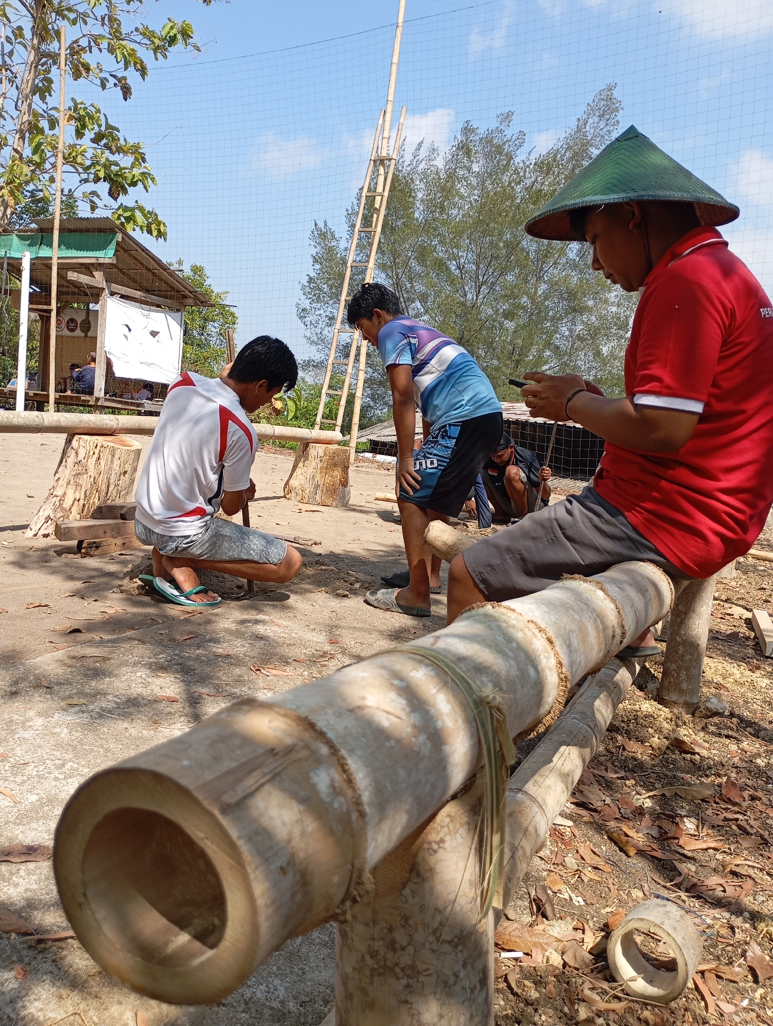 In Ketepung, Indonesia, on a sunny day, three individuals are engaged in an outdoor activity. One man, wearing a red shirt, grey shorts, and a conical green hat, sits on a thick bamboo log, looking at his phone. To his left, another man in a white and red shirt and grey shorts kneels on the ground, actively working on a piece of bamboo with a tool. In the background, a third man in a blue and white patterned shirt and black shorts is also present, and another person can be seen sitting inside a small wooden structure. The environment is dusty with scattered dry leaves, and bamboo poles and cut logs are visible throughout the scene. A net covers the area above, supported by bamboo poles, and a wooden ladder leans against some bamboo.