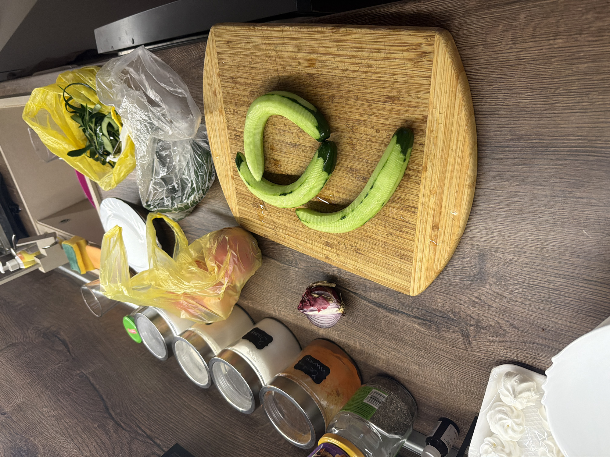 The image shows a cluttered kitchen counter. A wooden cutting board holds three cucumber pieces, two of which are curved. To the left of the board are a yellow bag filled with cucumber peelings, a clear plastic bag containing vegetables, and a white plate. Below, spices are stored in jars. Nearby are an onion, meringues, and more plates. No persons are visible. The location is Polski Trambesh, Bulgaria.