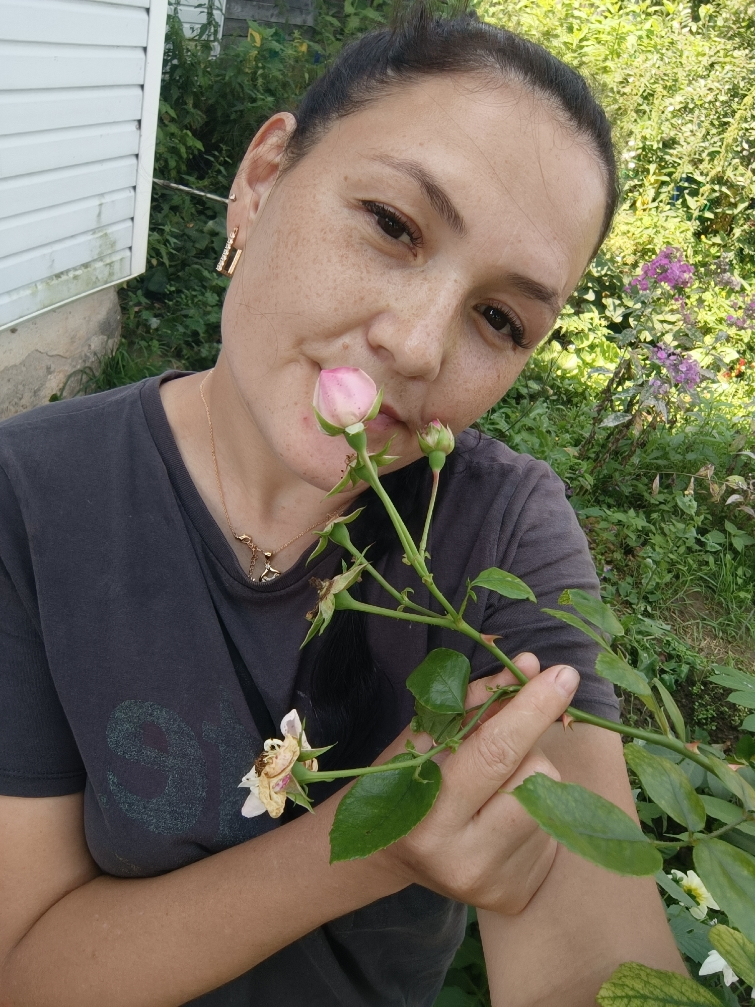 A medium shot photograph shows a woman in an outdoor setting, holding a rose stem with one pink bud and a wilting white flower. She is wearing a dark grey t-shirt and a gold necklace with a pendant. The background consists of lush green foliage and a section of a white, clapboard-sided building. The woman is looking towards the camera with a slight tilt of her head. The image does not contain enough information to determine the specific location within Karsha, Russia.
