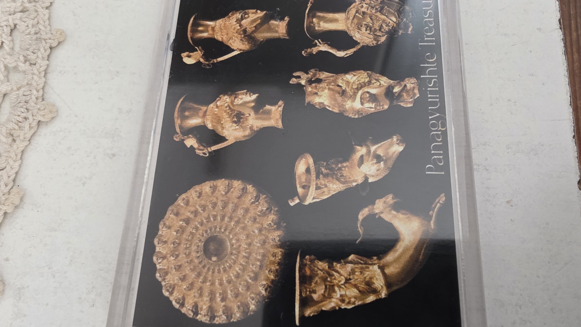 The image shows a photograph of a collection of golden artifacts, labeled "Panagyurishte Treasure," likely a reproduction or image of the original treasure. The objects include ornate drinking vessels or rhytons, some shaped like animal heads and others with human figures, and a flat circular dish with a repeating pattern. The image may be found in the city of Blagoevgrad, Bulgaria, due to the proximity of Panagyurishte.