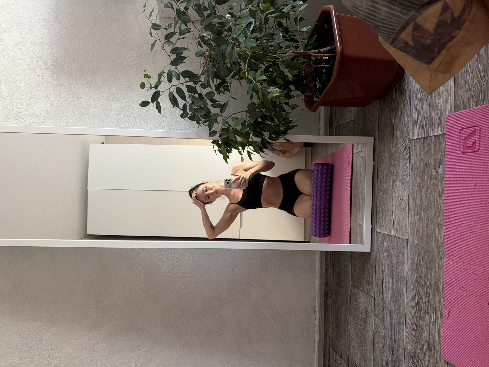 A slender woman in black sports attire stands on a pink yoga mat in what appears to be an indoor space. Her image is reflected in a full-length mirror with a white frame. She's holding a smartphone and appears to be taking a selfie. Behind the mirror, a large potted plant with lush green foliage is visible, suggesting an indoor environment. The floor is covered with gray wood-like tiles. A glimpse of another pink yoga mat is on the right side of the image. There is no evidence of the image being taken in Perm, Russia. The image lacks any geographical indicators to definitively place it in Perm or anywhere else.