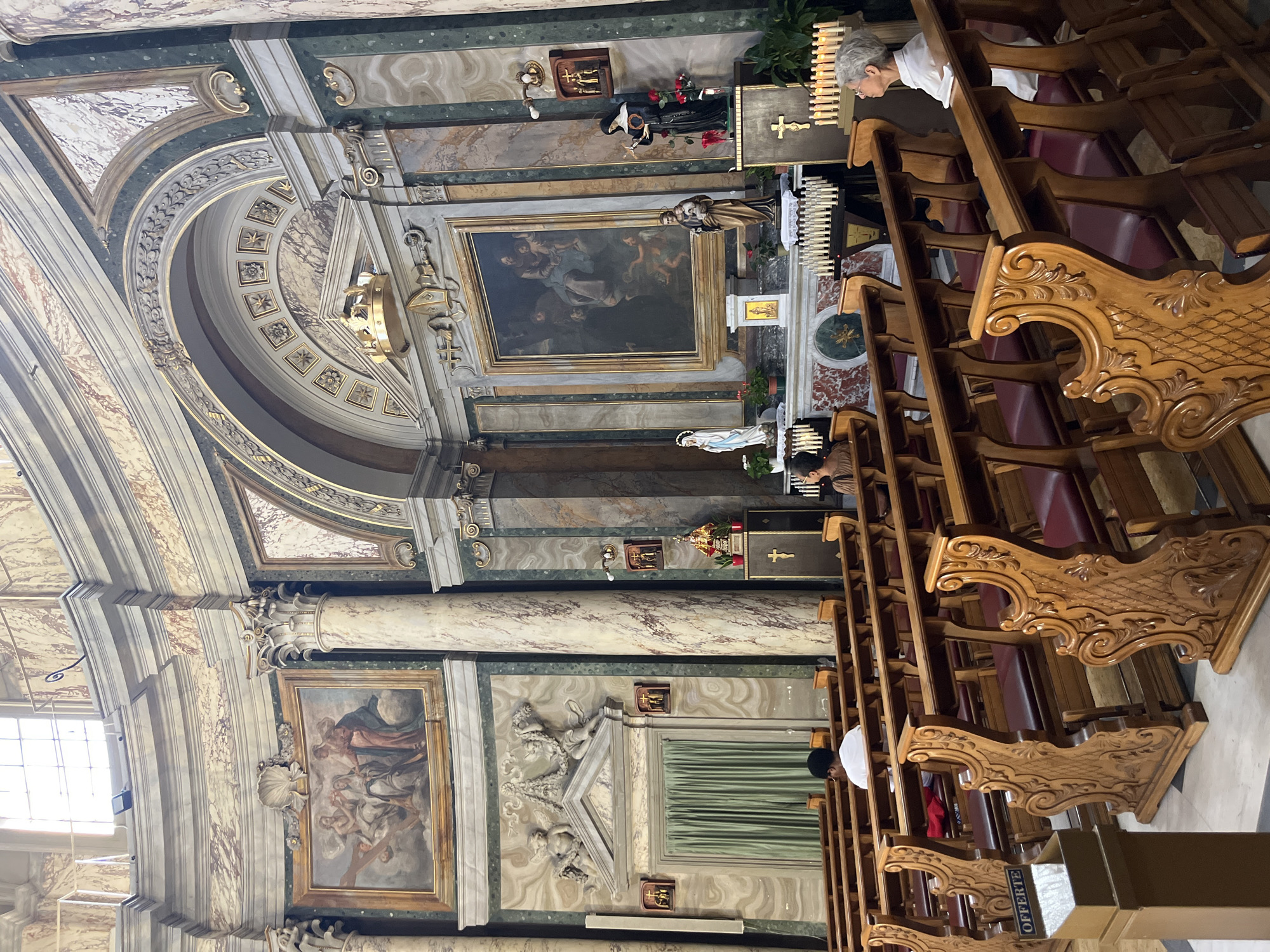 The image shows the interior of the Basilica of Saint Paul Outside the Walls in Rome, Italy. The photograph captures a view of the nave and the apse, with ornate marble columns and decorative frescoes adorning the walls and ceiling. Rows of wooden pews with red cushions are visible, occupied by a few individuals. In the apse, there is a large painting above the altar, flanked by statues and religious iconography. The lighting suggests it is daytime, with light filtering in from windows. The overall atmosphere is one of solemnity and architectural grandeur.