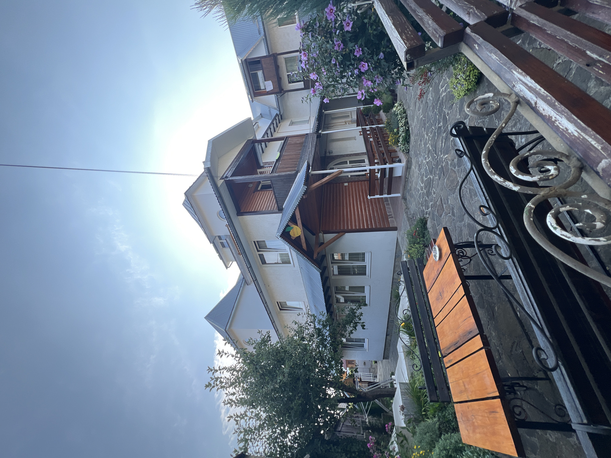 The image shows a courtyard area in front of a multi-story building, likely a guesthouse or hotel, in Yaremche, Ukraine. The building has white-painted walls and dark roofs, with wooden accents on balconies and porches. There is a tree on the left side of the frame, and a bush with purple flowers on the right. In the foreground, there is a wooden table with benches. The ground is paved with irregular stone tiles. The sky is visible at the top of the image, with the sun creating a bright flare. There are no people or animals present in the image.