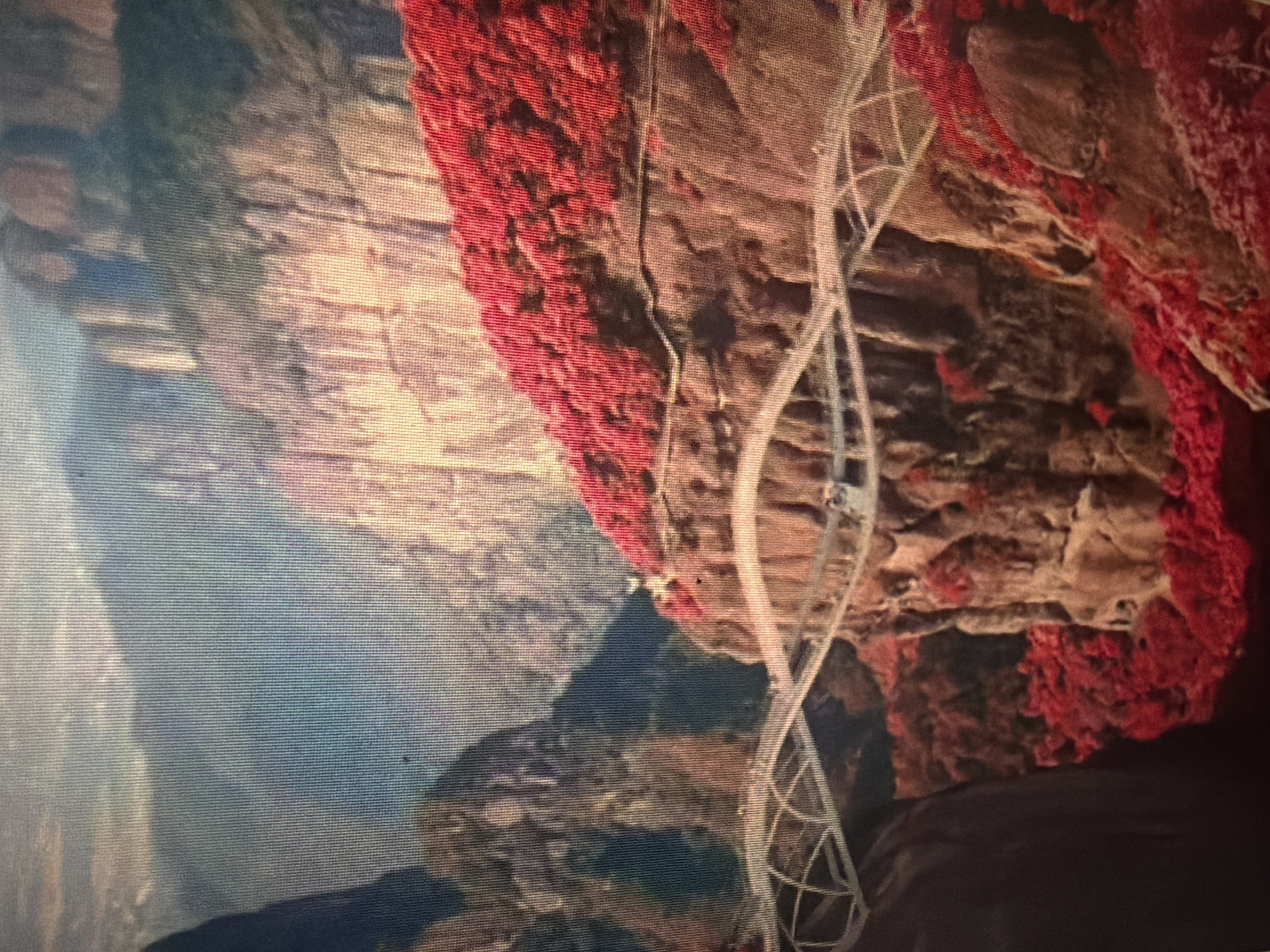 The image shows a wide, winding bridge suspended between two rocky cliffs. The cliffs are covered in dense, vibrant red foliage, contrasting with the muted, bluish-gray of the distant mountains. The sky above is a pale blue with scattered white clouds. The bridge appears to be made of glass and metal, with a series of connected geometric panels forming its structure. It curves and undulates as it spans the chasm, giving the impression of a delicate, almost ethereal pathway. The overall scene evokes a sense of awe and wonder, highlighting the dramatic natural landscape and the daring architectural feat. The location is identified as Baan Mae Nam, Thailand, though no specific landmarks within the city are discernible. The image does not depict any people or overt actions, focusing solely on the impressive natural and man-made structures.