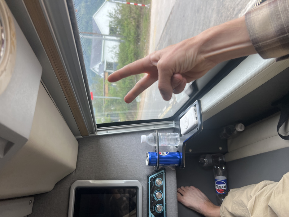 The image depicts the interior of a passenger vehicle, likely a sleeper bus, looking outwards through a window onto a roadside environment in M'Drắk, Vietnam.
**Interior:**
On the left, a bare human left foot is visible, resting on a beige upholstered seat or footrest. Further up and left, a small, dark-screened monitor with a white bezel is embedded in a grey fabric wall. Below the monitor, a control panel features two circular dials (one blue, one silver) and multiple smaller buttons, with partially visible text including "Light" and "USB." A black metal console extends from the wall towards the window, featuring two circular cup holders. The left holder contains a blue Pepsi soda can; the right holder contains a clear plastic bottle of water. To the right of the cup holders, on an integrated flat tray, rests a white portable power bank with a white circular pop socket attached. Below the console, on the vehicle floor, two empty plastic Pepsi bottles are present. A black strap, possibly from a bag, is also visible. The vehicle interior features grey fabric wall coverings and beige/off-white seating and trim.
**Human Element:**
In the right foreground, a pale-skinned human right arm, wearing a sleeve of a brown and white plaid shirt, is extended towards the window. The hand is performing a "peace" or "V for victory" gesture with the index and middle fingers extended upwards, while the ring and pinky fingers are curled into the palm. Visible veins are present on the forearm.
**Exterior Environment (M'Drắk, Vietnam):**
Beyond the window, an asphalt road with a yellow double line marking is visible. The immediate roadside features green grass and vegetation. In the mid-ground, a white gabled house with a dark roof stands amidst greenery, alongside a smaller, partially obscured structure. A red rectangular sign or marker is visible near the house, and a white and red striped marker pole stands to the far right. The background consists of lush green hills or mountains, suggesting a rural or semi-rural setting. The lighting appears bright and overcast.