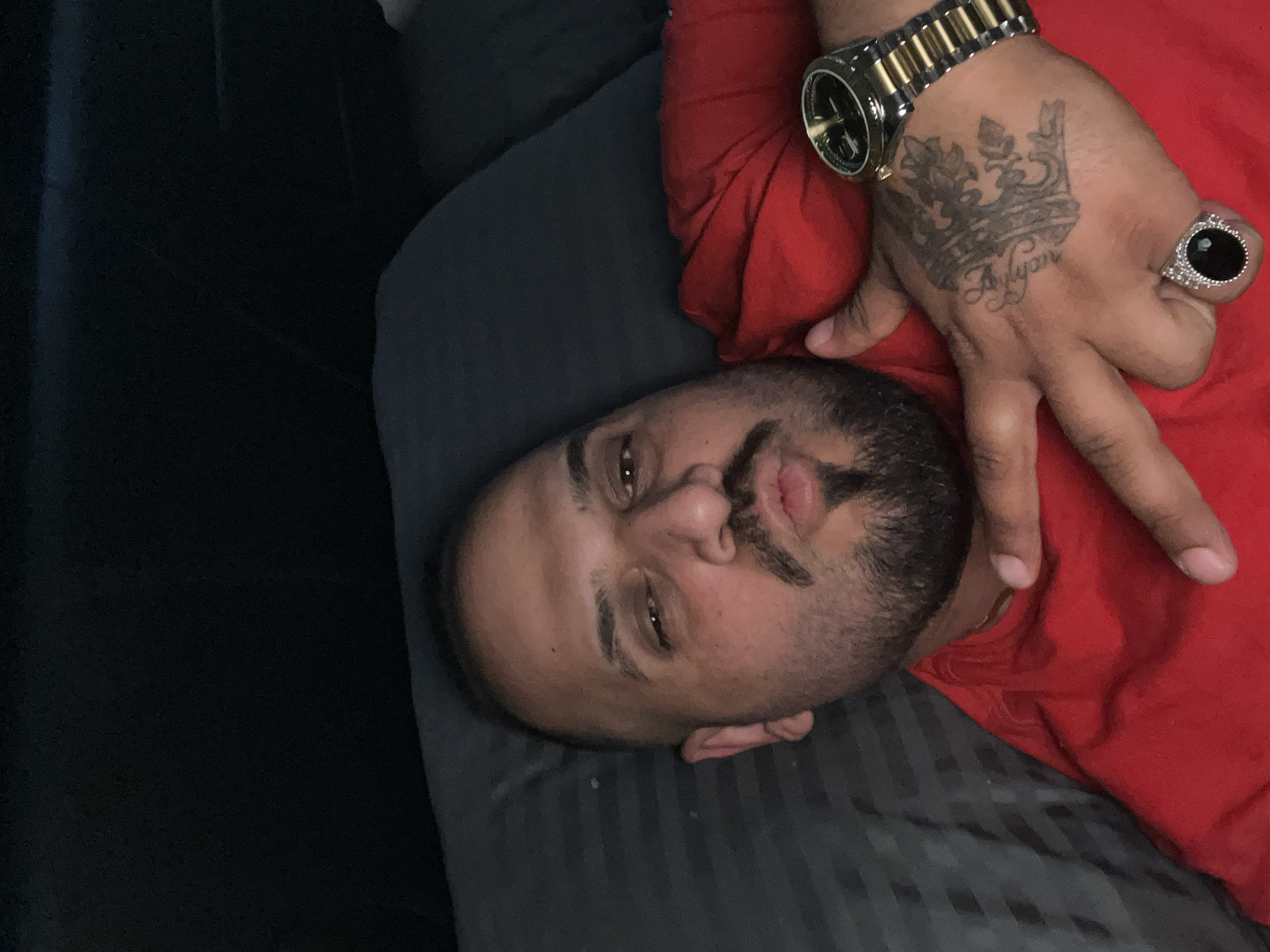 The image displays a man of Middle Eastern or South Asian descent lying down, with his head turned to the left and looking towards the camera. The photograph is oriented vertically, showing the man's head and upper torso. He has a full beard and mustache, and his expression is neutral with slightly pursed lips.
He is wearing a red t-shirt or garment. On his right hand, which is visible in the upper right portion of the image, he wears a prominent gold-and-silver wristwatch with a black face. He also has a large silver-toned ring on his ring finger, featuring a substantial black oval stone surrounded by smaller clear stones. A distinctive tattoo of a crown is visible on the back of his hand, with what appears to be the name "Rayan" (or similar) tattooed beneath it in a script font. His hand is slightly raised, with fingers splayed.
The man is resting on a dark grey or black surface, possibly a bed or couch, which has subtle stripes. The background is predominantly dark, suggesting an indoor setting with low or ambient lighting. No specific location cues are visible beyond the bedding.