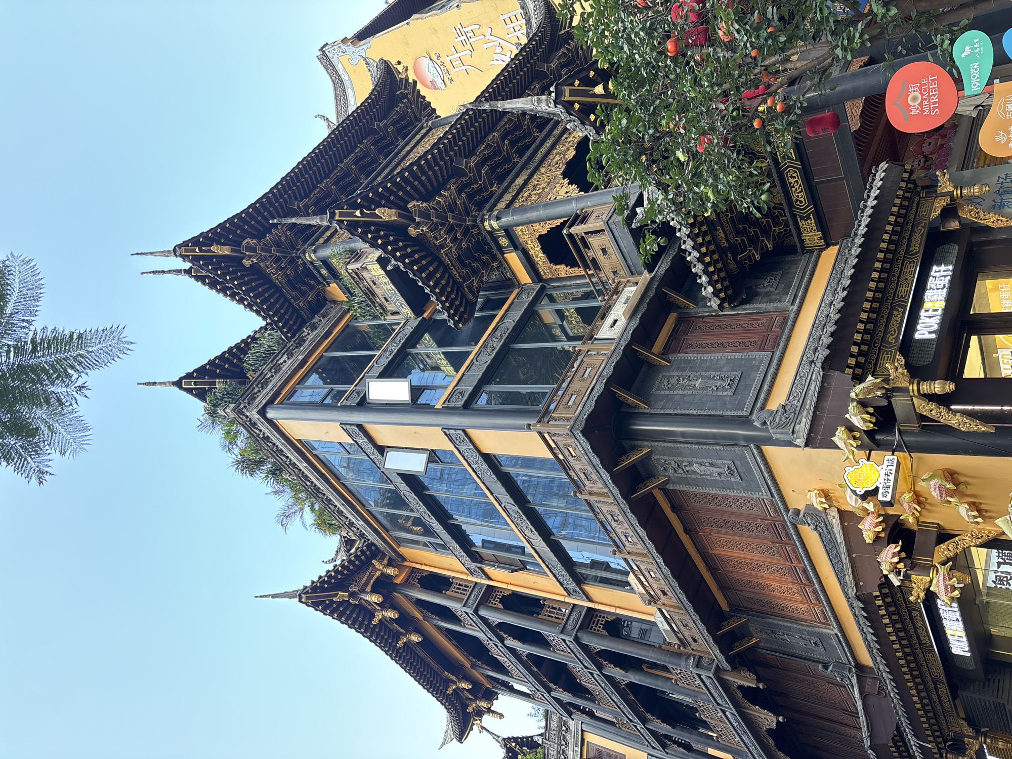 The image shows a multi-story building with traditional East Asian architecture in Yuzhong District, China. The building features tiered, dark, ornate roofs with golden trim and decorative elements. The upper floors have large glass windows reflecting the sky and surrounding buildings. The lower sections of the building display dark wooden latticework and ornate carvings. A tree with red fruit and lanterns is visible on the right side of the image, with signage in Chinese characters. The sky is clear and light blue.
