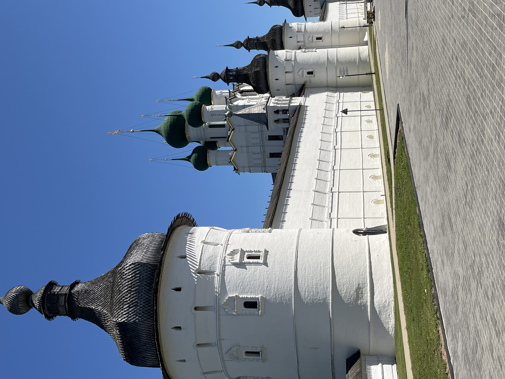 The image shows a bright, sunny day with a clear blue sky over the city of Novo-Nikolskoye, Russia. The scene is dominated by a large, white, fortress-like complex with several towers. The main tower in the foreground is round and white, topped with a dark, onion-shaped wooden roof. Adjacent to it, a long white wall with arched openings runs horizontally across the image, connecting to other round towers. In the background, behind the wall, several white church buildings rise with distinctive green onion domes, some topped with golden crosses. The ground in front of the complex is paved with gray interlocking stones, and a strip of green grass runs along the edge of the wall. A lamppost and a bench are visible in the distance on the paved area. The architectural style suggests a historic religious or defensive structure. There are no people visible in the image.