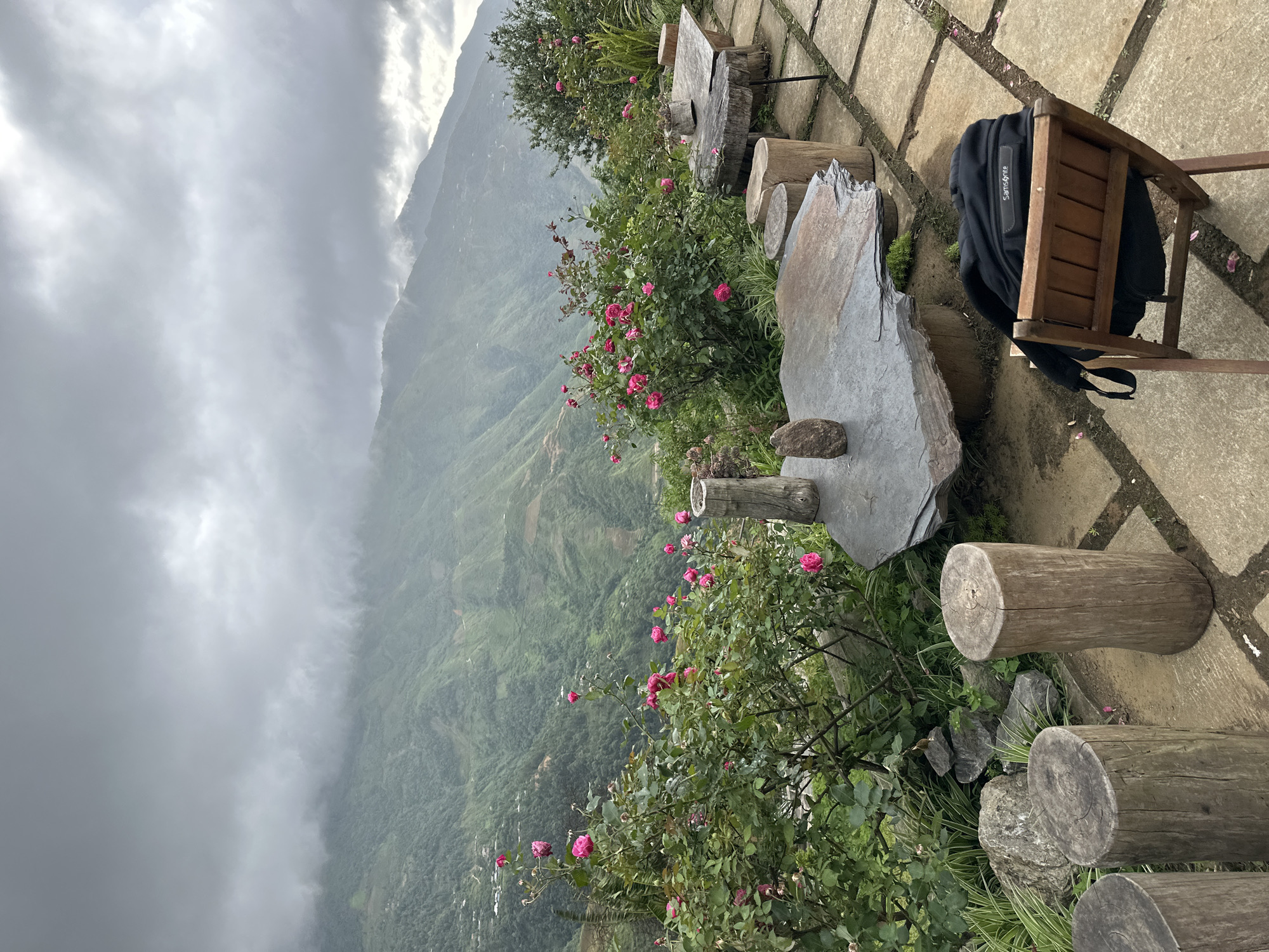 The image captures a scenic view from a cafe or guesthouse terrace in Ta Xua, Vietnam, overlooking a lush, mountainous landscape shrouded in mist and clouds. The foreground features a rustic seating area with several rough-hewn wooden log stools and two large, flat slate tables. A black Samsonite backpack rests on a simple wooden chair placed on a paved stone pathway. Pink roses bloom in abundance in bushes bordering the terrace, adding a touch of color against the muted greens of the mountains and the gray sky. The mountainous terrain is densely forested, with evidence of terraced farming visible in the distance. The overall atmosphere is one of tranquility and natural beauty, enhanced by the dramatic cloud formations.