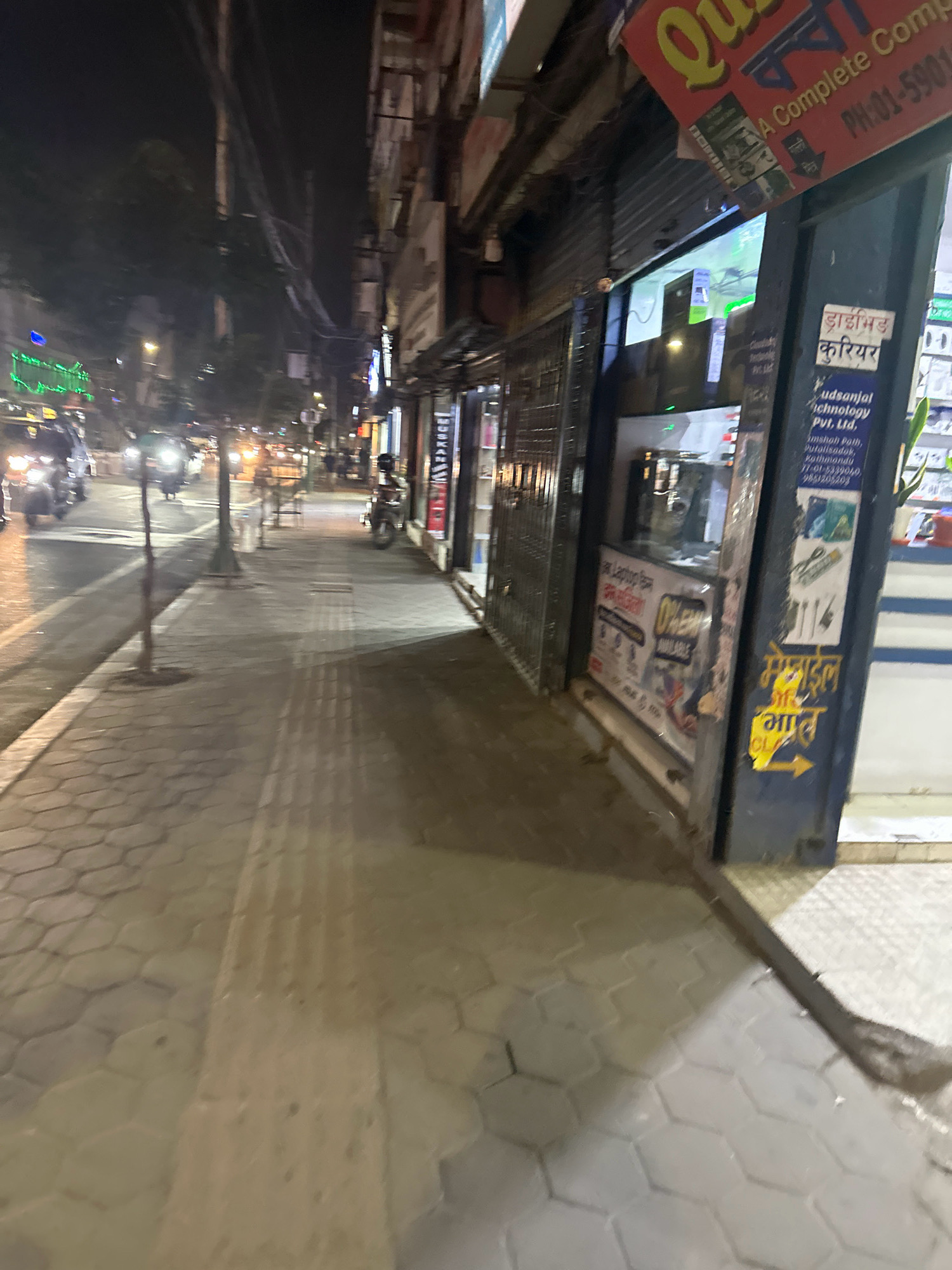 This image depicts a street scene at night in Kathmandu Metropolitan City, Nepal. A pedestrian sidewalk, paved with hexagonal tiles, runs along the right side of the frame. A tree grows from the sidewalk, casting a shadow. The street, with traffic lights and oncoming vehicles, is visible on the left. Shops line the sidewalk on the right, with some displaying illuminated signs and product advertisements. One shop's signage includes text in Nepali script and English, indicating it's a courier service and technology provider. The overall atmosphere is that of a busy urban street at night.