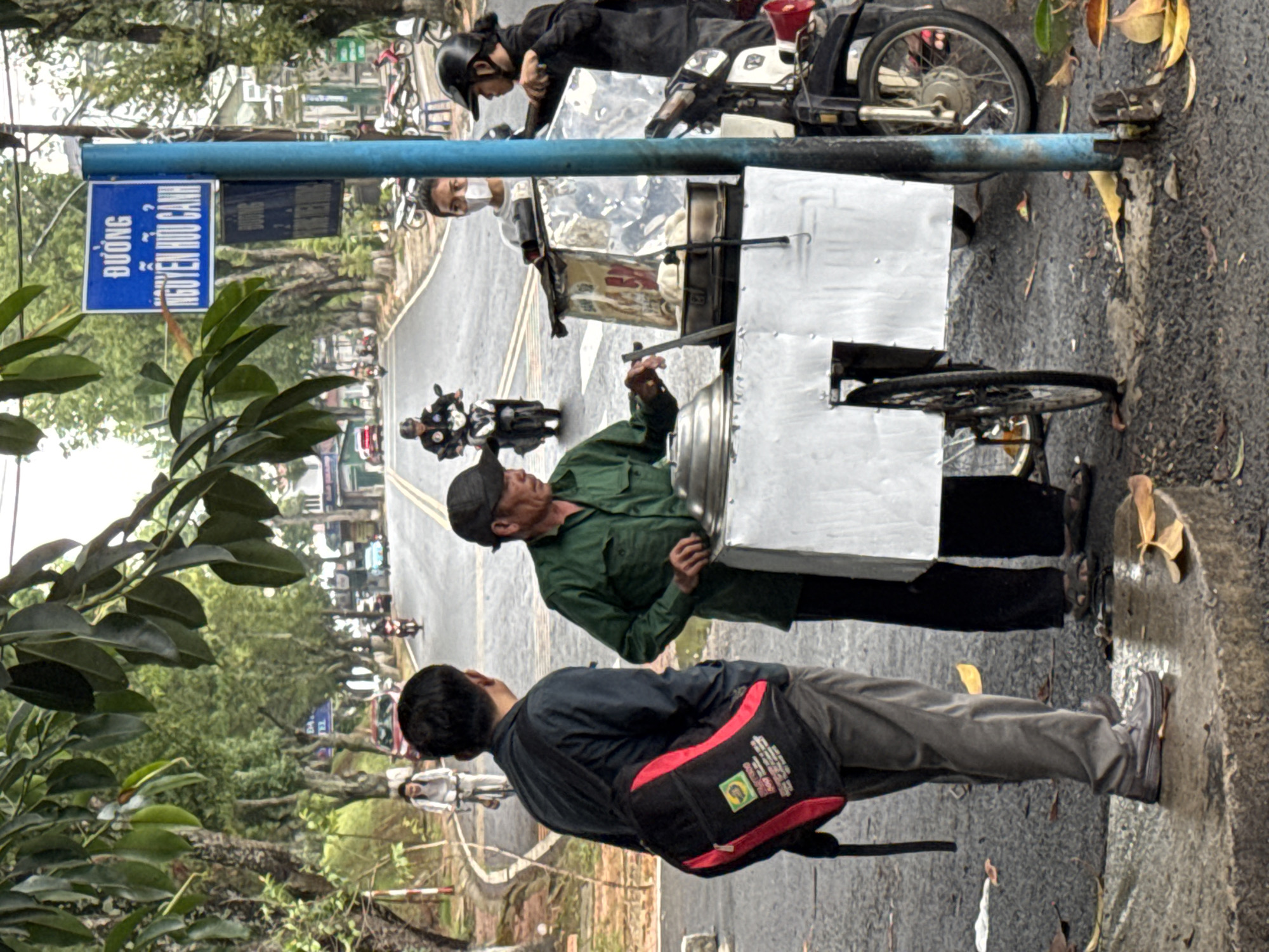 This image depicts a bustling street scene in Lâm Viên - Đà Lạt, Vietnam, centered around a street food vendor. The setting is an outdoor area adjacent to an asphalt road with visible yellow double lines, bordered by trees and lush green foliage.
In the mid-ground, a male street vendor, dressed in a green shirt and a dark cap, is actively stirring the contents of a large pot situated on a mobile metal cart. He appears focused on preparing or serving food. To his left, seen from behind, a person wearing a dark jacket, light pants, and a black and red backpack observes the vendor. Another individual in a white shirt is partially visible behind the vendor's cart. On the right, a person on a motorbike, clad in a black jacket and helmet, looks towards the vendor, suggesting an interaction or transaction. Further down the tree-lined street, other motorbikes and pedestrians can be seen, contributing to the everyday street activity.
A prominent blue street sign, attached to a blue pole, features clear Vietnamese text: "ĐƯỜNG" (Street) and "NGUYỄN HỮU CẢNH", identifying the street as Nguyễn Hữu Cảnh Street. The lighting indicates it is daytime, possibly under an overcast sky or during the softer light of early morning or late afternoon, as shadows are diffused. The overall atmosphere is that of a typical, active street in Vietnam. The backpack worn by the foreground person has a distinctive red and black design, along with some unreadable white text and a small square logo.