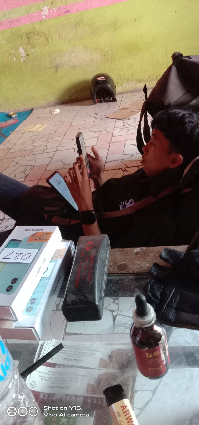 The image displays an indoor scene in Kandangjati Kulon, Indonesia, depicting a male individual resting in a reclining position on a tiled floor, engaged with multiple electronic devices.
The central figure is a young male with short dark hair and a dark complexion, wearing a black short-sleeved top with white lettering (partially visible "IS") and a black smartwatch on his left wrist. He is propped against a dark backpack, holding one smartphone with both hands while a second illuminated smartphone rests on his body or the bag.
In the immediate foreground, a glass-top table or surface is visible, holding several items. These include two stacked white boxes for "Samsung Galaxy A06" mobile phones, with the top box displaying "25W GRATIS" and "027". Next to these are a black rectangular object with a red graphic, a pair of black gloves, and a small dark glass bottle with a dropper cap containing a reddish-brown liquid. A light-colored cigarette lighter with "ARWA" text and papers featuring Indonesian warnings (including "PERINGATAN" and "FILTER CIGARETTES" with a "21+" age restriction) are also present. A transparent plastic bottle is partially visible on the far left.
The environment consists of a light-colored, patterned tiled floor and a worn lime-green wall with horizontal pink stripes and visible peeling paint. On the floor in the background, a black motorcycle helmet with "IRON M" branding is positioned near the wall. A light blue mat or board, a crumpled white paper, and loose cardboard pieces are also on the floor.