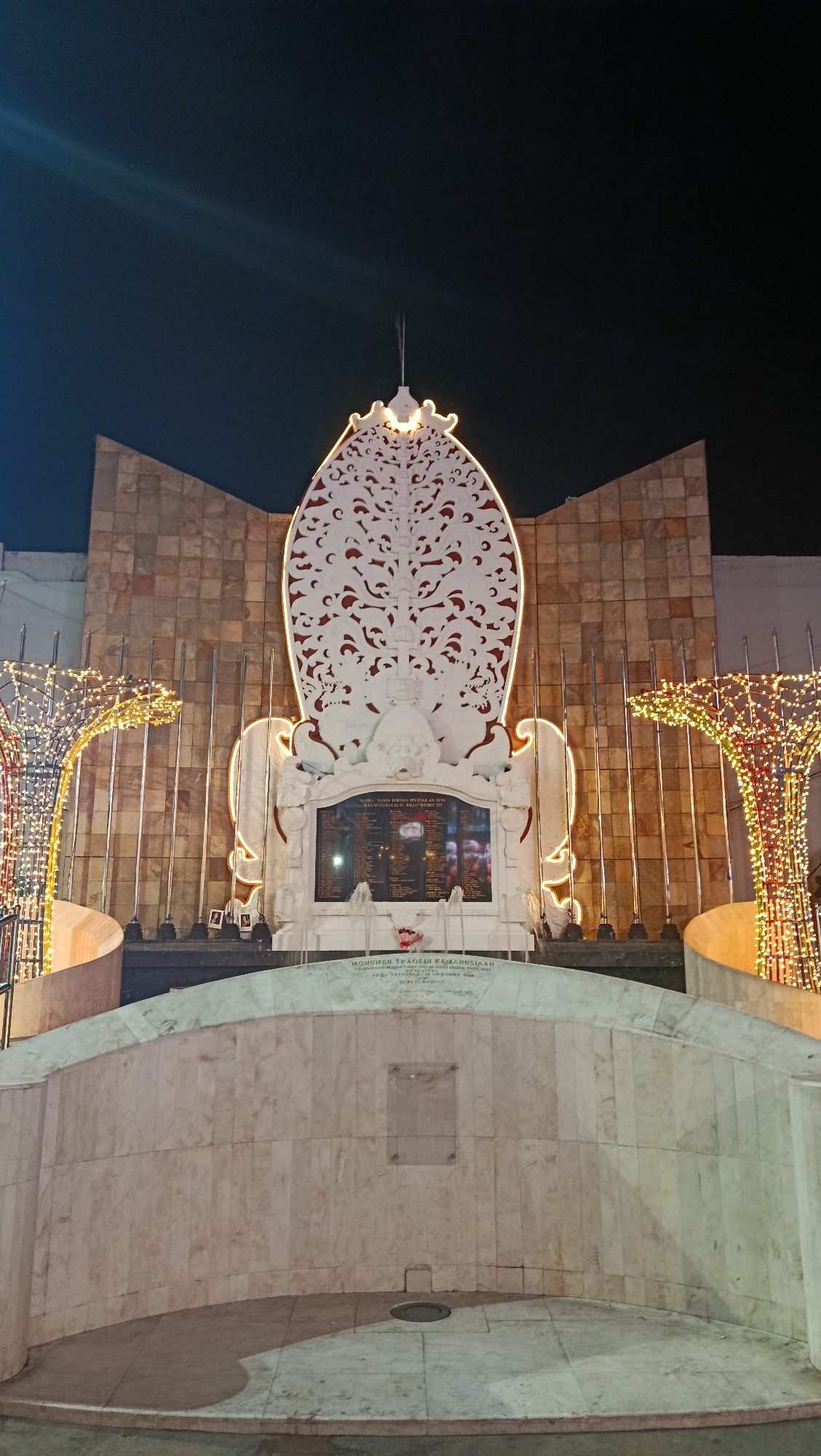 The photograph captures the Monumen Peringatan Bom Bali I (Bali Bombing Memorial) in Kuta, Indonesia, at night. The memorial features a central white stone structure with intricate carvings and a list of victims' names inscribed on a dark plaque. The structure is illuminated with yellow lights around its perimeter, and two large, decorative trees covered in yellow and red lights stand on either side. The background consists of a stone-tiled wall and flags on poles. In the foreground, a curved marble structure with a circular drain in the center is visible. The sky is dark, indicating it is nighttime. There are no people visible in the scene.