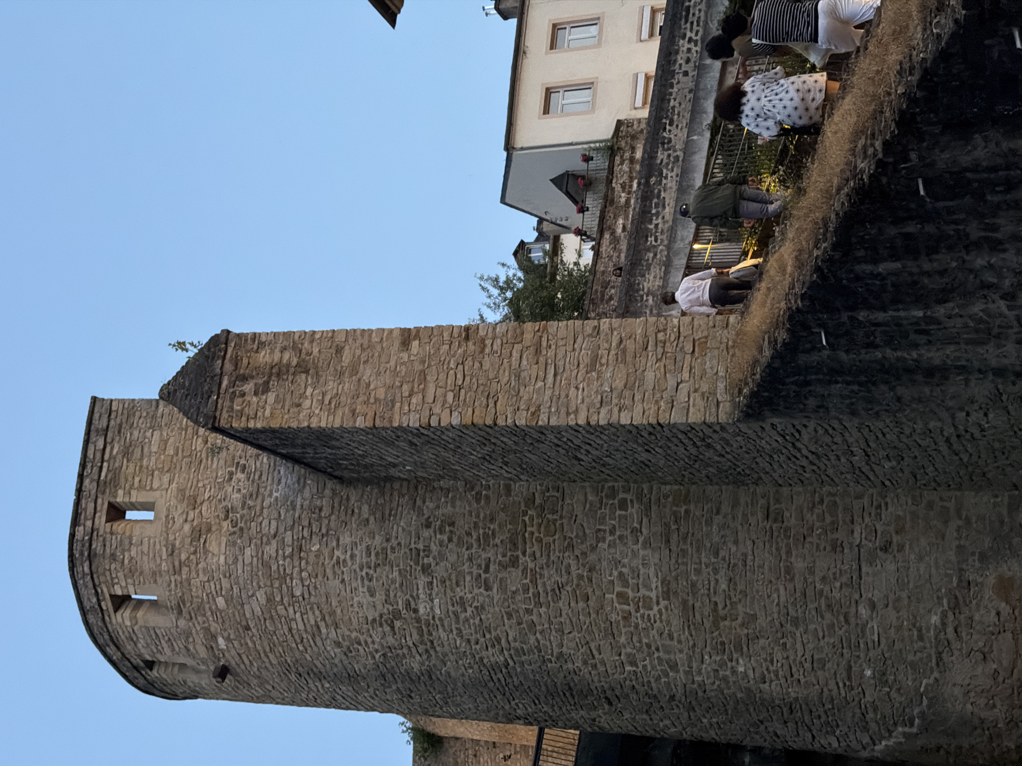 This image depicts a group of people ascending a stone staircase in front of a fortified tower in Luxembourg City, Luxembourg. The tower is constructed of large, rough-hewn stones and has a curved facade with openings at the top. Adjacent to the tower is a section of wall, also made of stone. Several individuals are visible walking up the stairs, including a person in a white shirt and gray pants, a person in a green jacket and jeans, and two individuals further up the stairs, one in a white dress with a floral pattern and another in a striped shirt and white pants. The background shows buildings with windows and balconies, nestled against a clear blue sky. The scene suggests a historical or tourist location, possibly part of the Bock Casemates or a similar ancient fortification within Luxembourg City.