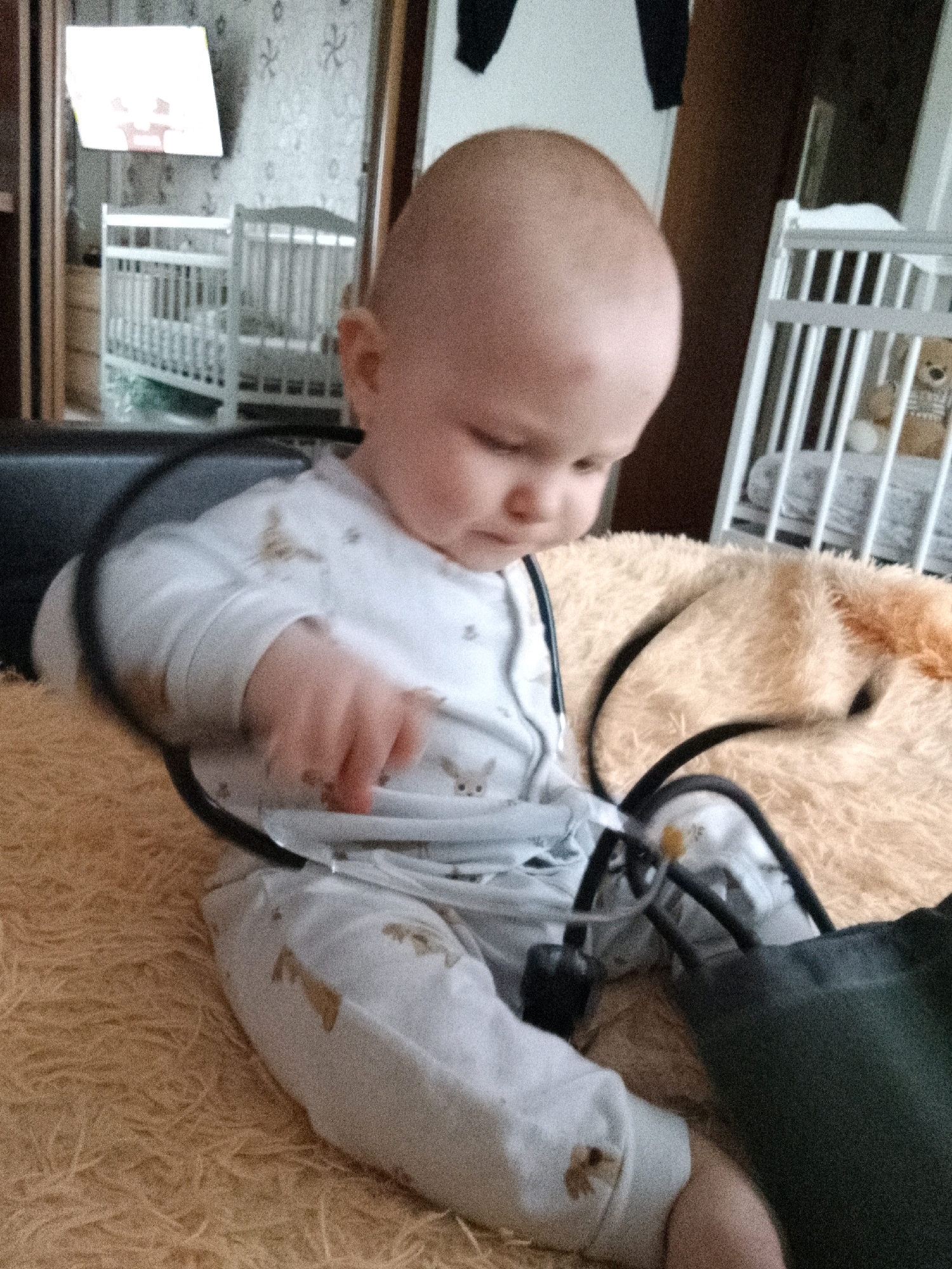 A baby in a light-colored onesie with animal prints is sitting on a fluffy, tan-colored surface. The baby is facing to the left and appears to be holding a stethoscope. In the background, there are two white cribs, one behind the other. The cribs appear to be in a bedroom or nursery. To the left of the cribs, there is a television mounted on the wall displaying a colorful image. The overall impression is of a domestic setting, likely a home in the city of Minsk, Belarus.