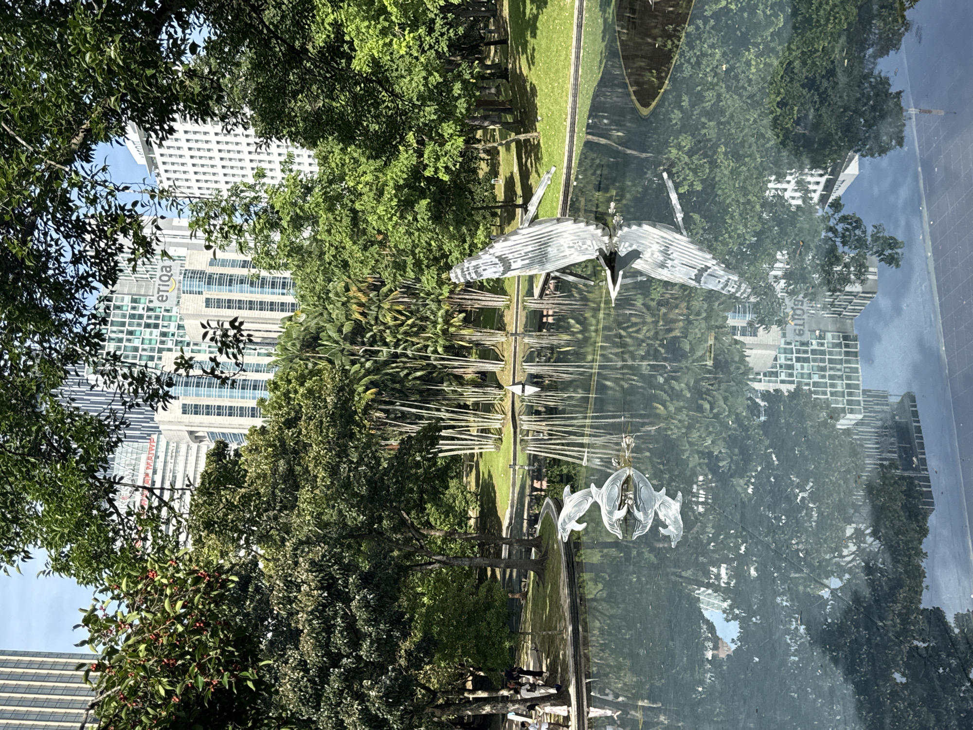 The image shows a view in Kuala Lumpur, Malaysia, possibly a reflection in glass or water, featuring a park-like setting with trees and buildings. There's a building with the word "etiqa" visible. A metallic sculpture of a bird or similar winged creature is prominently displayed. The scene includes elements of both urban architecture and natural green spaces.