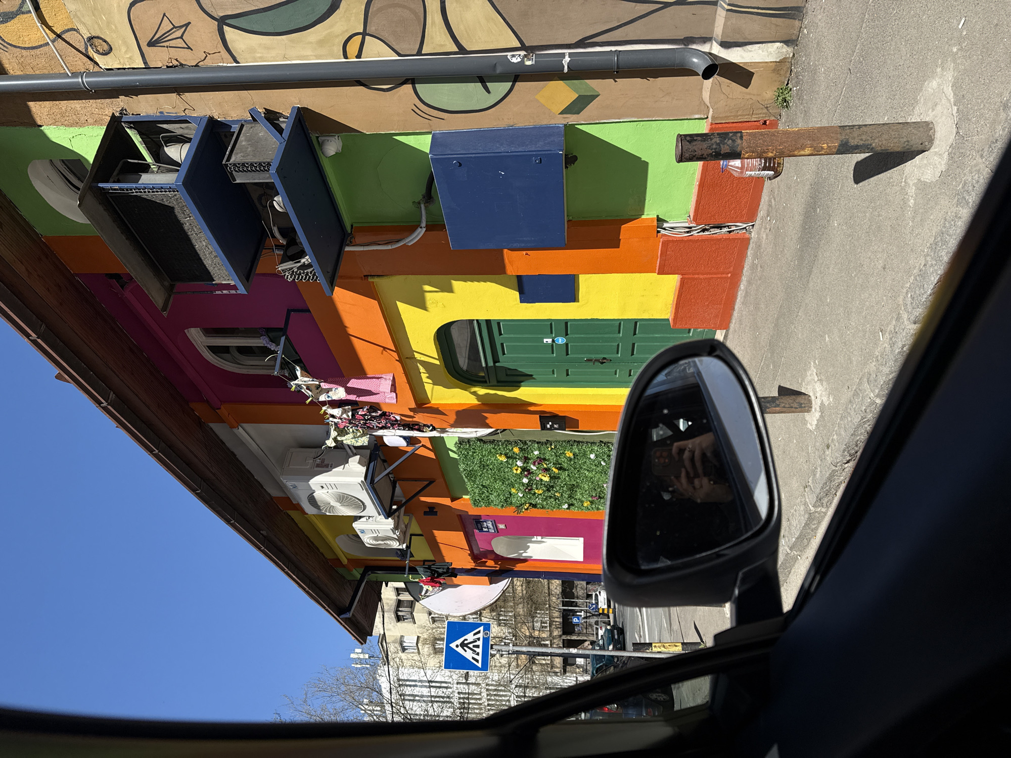 Captured from inside a car, the image depicts a colorful building in Belgrade, Serbia. The building's facade is painted in horizontal stripes of green, yellow, orange, red, and pink. A green door is visible. Various external fixtures, including air conditioning units, are attached to the building. A pedestrian crossing sign is seen in the background. The car's side mirror reflects the photographer holding a mobile device. The sky is clear and blue.