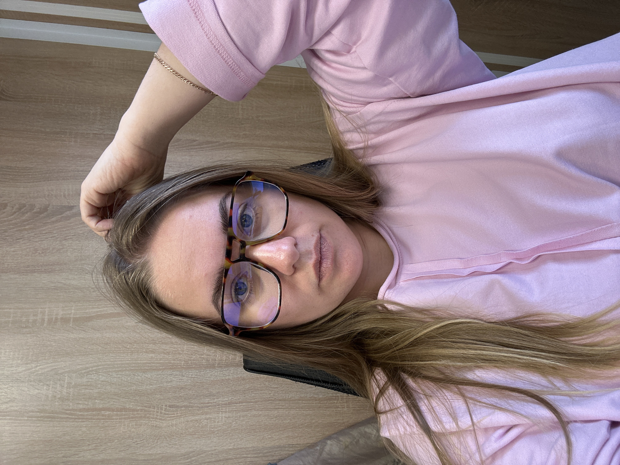 A woman with light brown hair and blue eyes wearing tortoiseshell glasses and a pink shirt is lying down. She is wearing a gold bracelet on her left wrist. The background appears to be a light-colored wooden surface. The location in the city of Ural'skoe, Russia, is not discernible from the image.